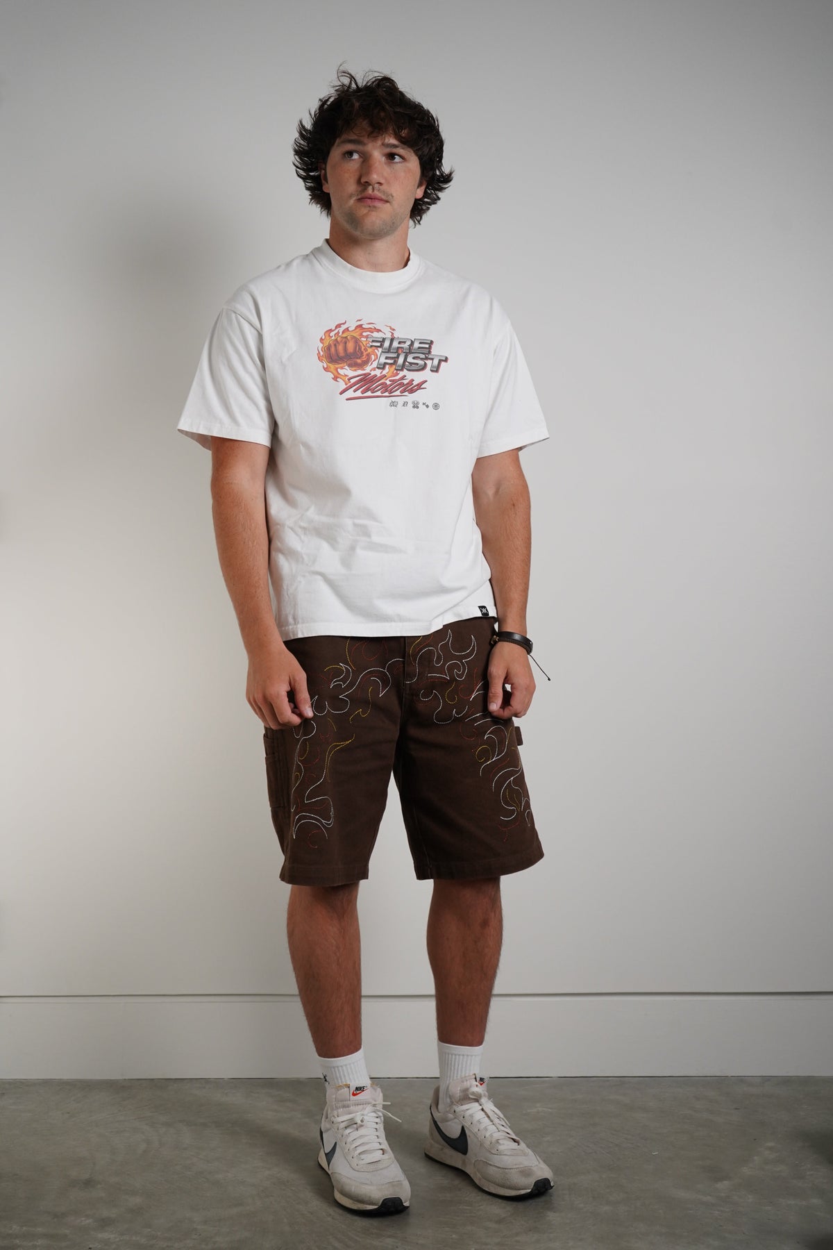 A man standing, wearing a white shirt and brown shorts, with white tennis shoes on concrete floor and white background.