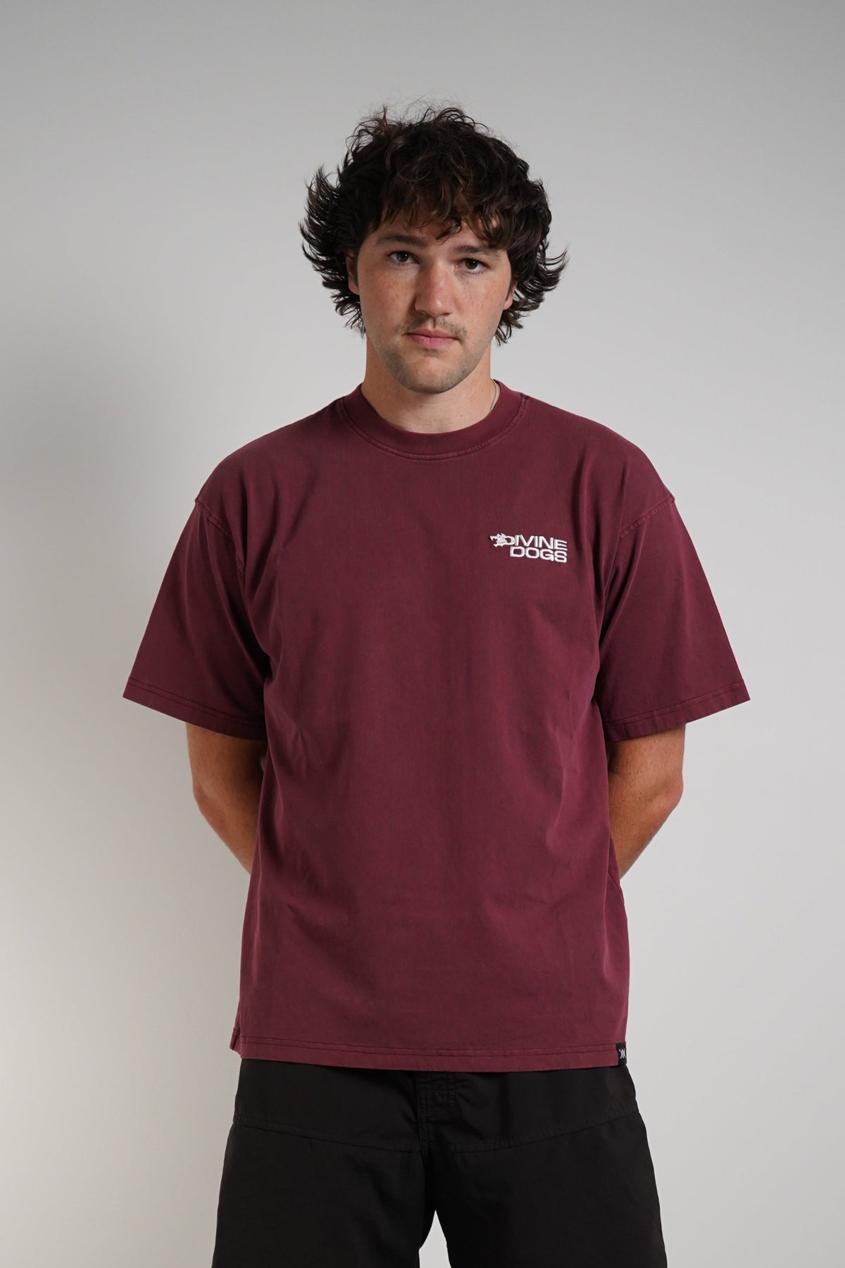A man wearing a burgundy t-shirt with a small white chest logo, black shorts, standing against a plain light gray wall.