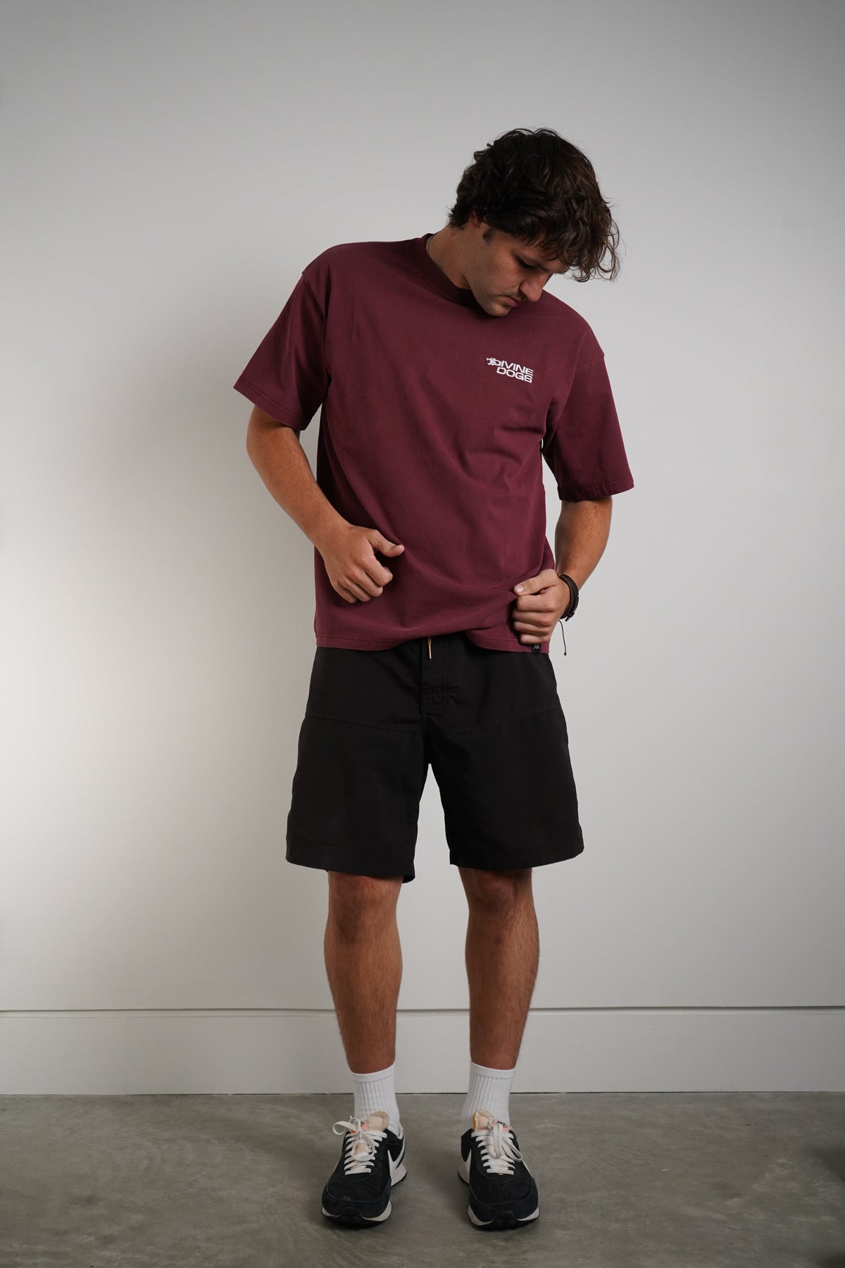 A man wearing a burgundy t-shirt with a small white logo on the left chest, black shorts, white socks, and black sneakers, standing against a plain light gray wall.