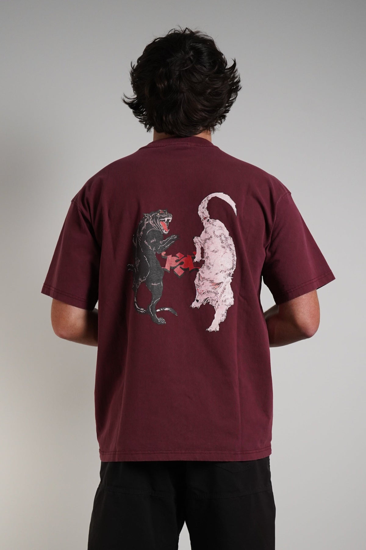A man viewed from behind wearing a maroon t-shirt with a graphic of a black dog and a white dog facing each other, paired with black shorts, standing against a plain gray wall.