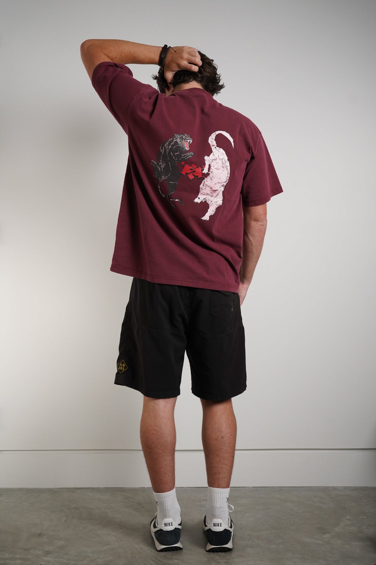 A man stands with his back to the camera wearing a maroon t-shirt with a graphic print of a black bear and a white cat, paired with black athletic shorts, white socks, and sneakers.