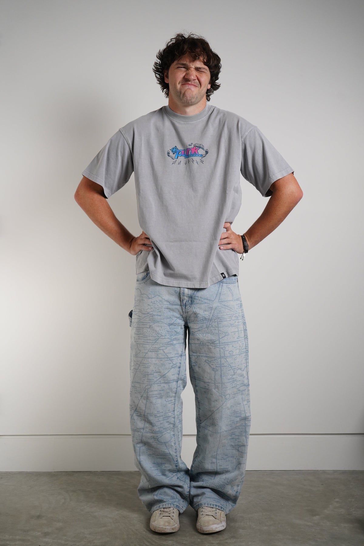 A man wearing a light gray crewneck t-shirt with a colorful graphic on the chest and light blue denim jeans, standing against a plain light gray wall.