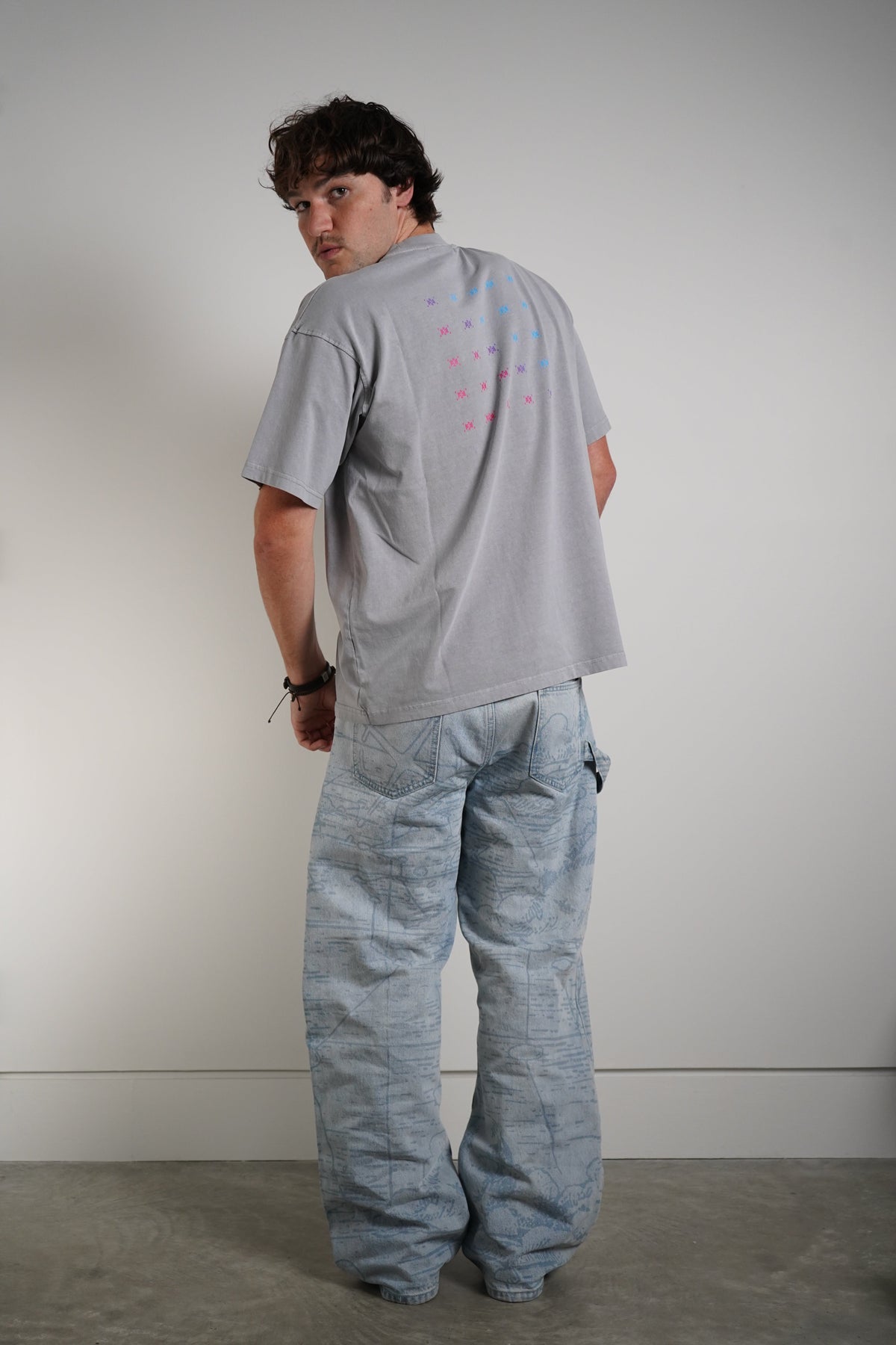 A man wearing a light gray oversized t-shirt with a colorful graphic print on the back and light blue wide-leg denim pants, standing against a plain light gray wall.