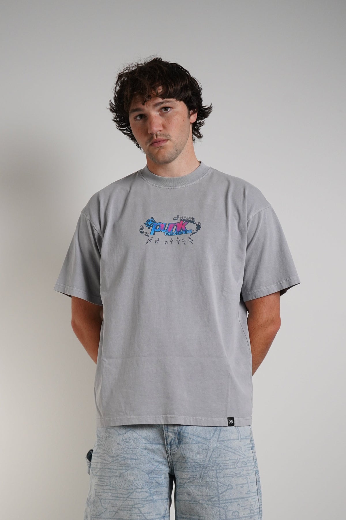 A man stands against a plain light gray wall wearing a light gray t-shirt with a colorful graphic on the chest, light blue marled sweatpants, and beige sneakers.