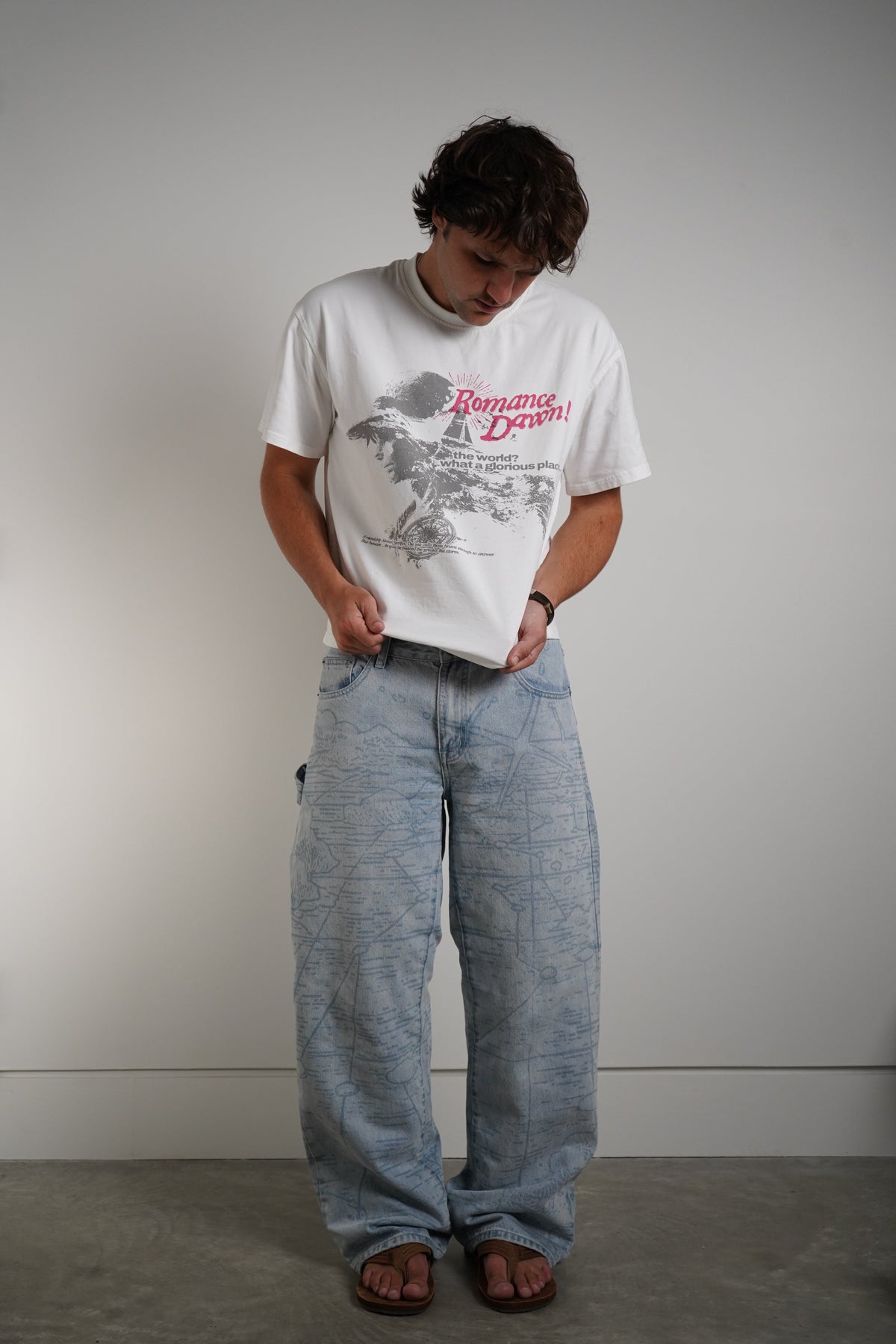 A man wearing a white graphic t-shirt with a red and gray print, paired with light-wash wide-leg jeans and brown sandals, standing against a plain gray wall.