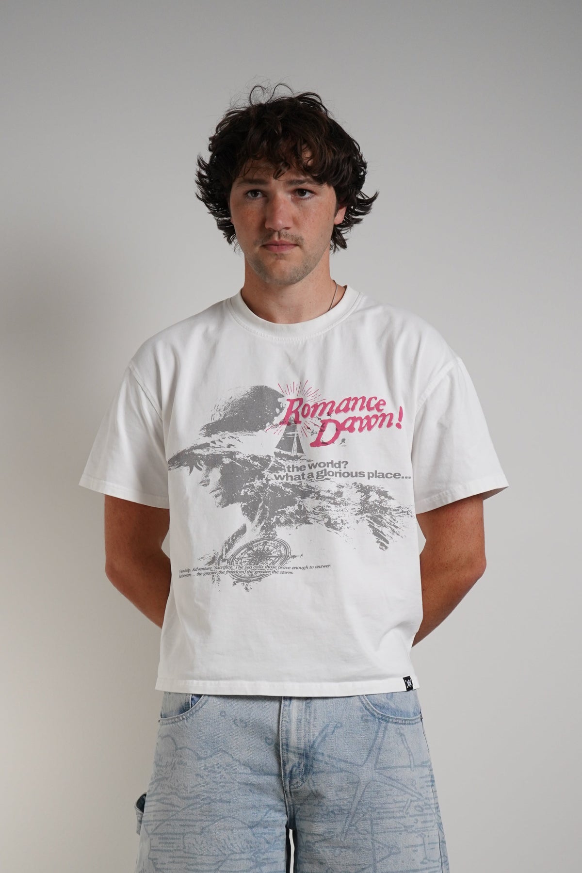 A man stands against a plain light gray wall wearing a white graphic T-shirt with a red and gray print, loose light-wash blue jeans, and brown sandals.