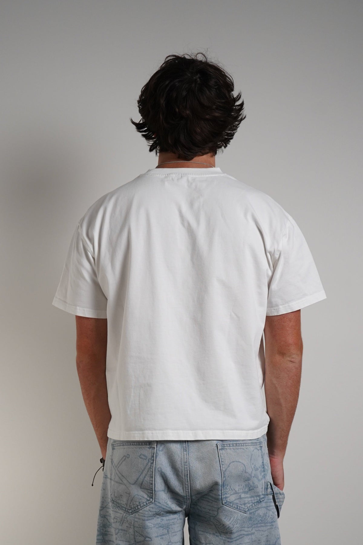 A man viewed from behind wearing a white short-sleeve t-shirt and light blue wide-leg linen trousers, standing against a neutral gray wall.