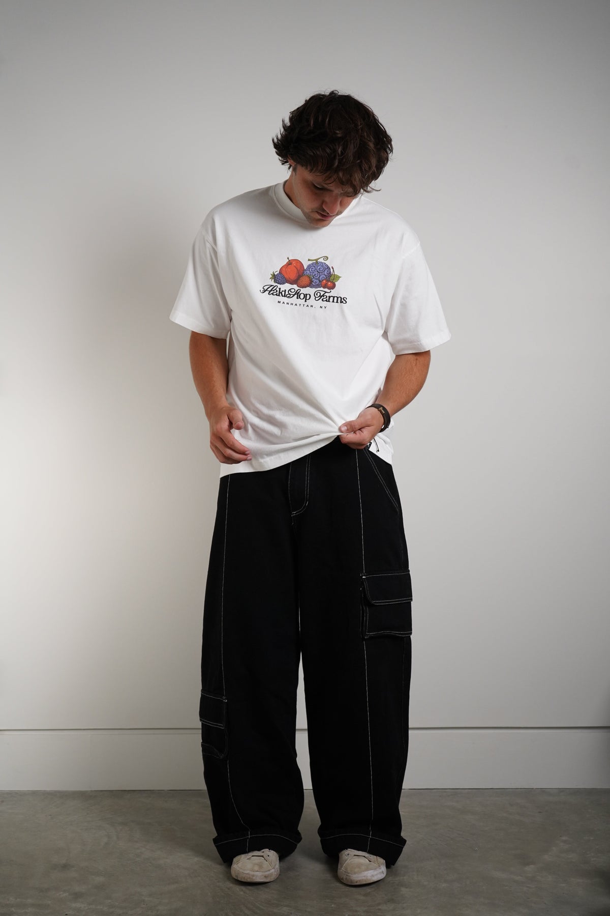 A person wearing a white graphic t-shirt and black wide-leg cargo pants, standing against a plain light gray wall.