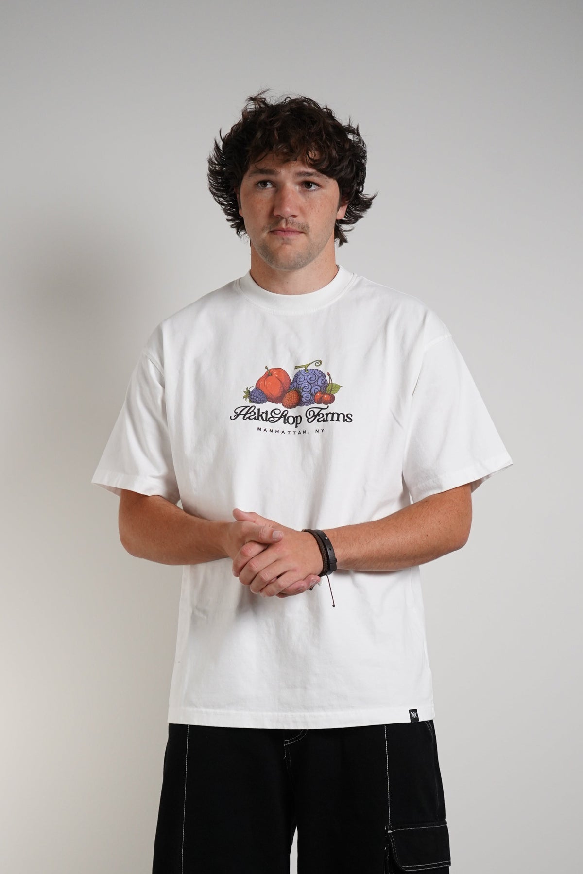 A man stands against a plain light gray wall wearing a white graphic T-shirt with a colorful fruit illustration and the text 'Hakistop Farms' on the chest, paired with loose black cargo pants and light-colored sneakers.