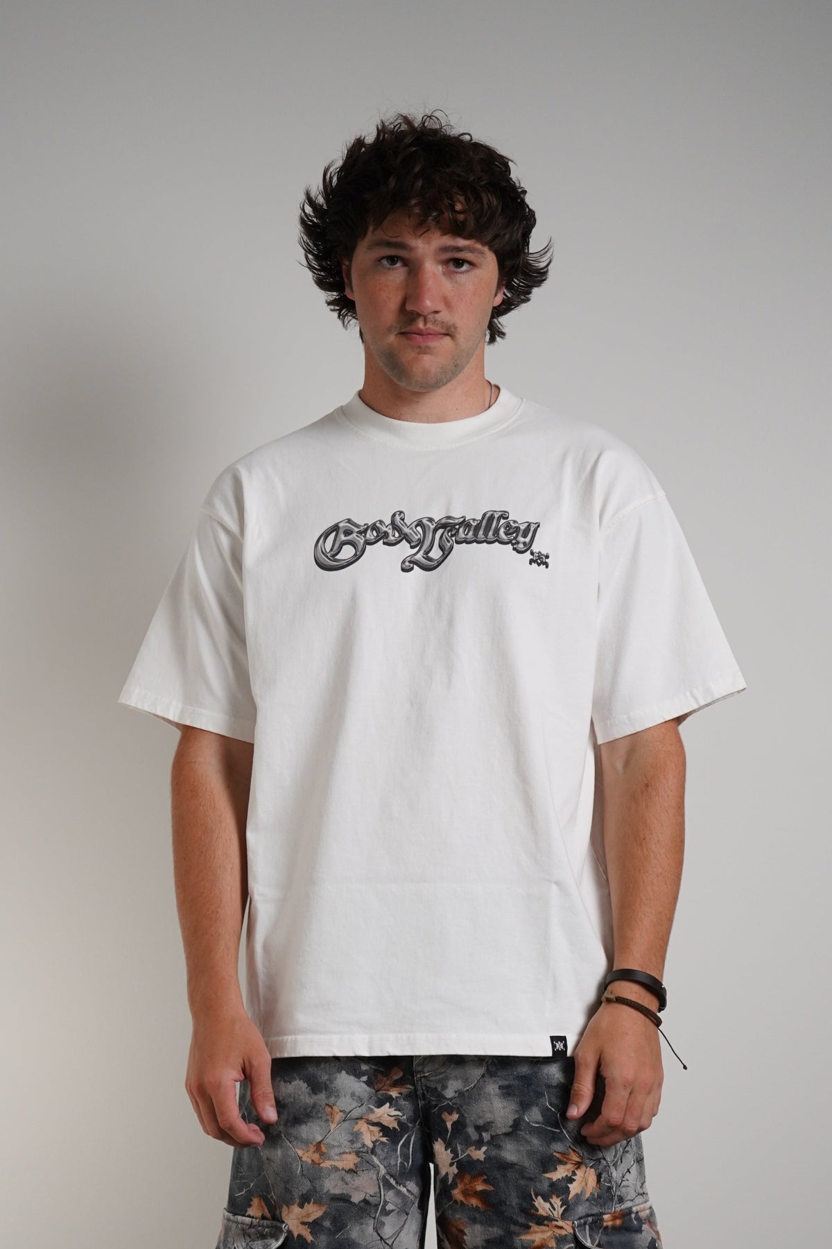 A man stands against a light gray studio wall wearing a white graphic T-shirt with a black script logo across the chest, loose dark gray cargo pants with orange distressed patches, and black low-top sneakers.