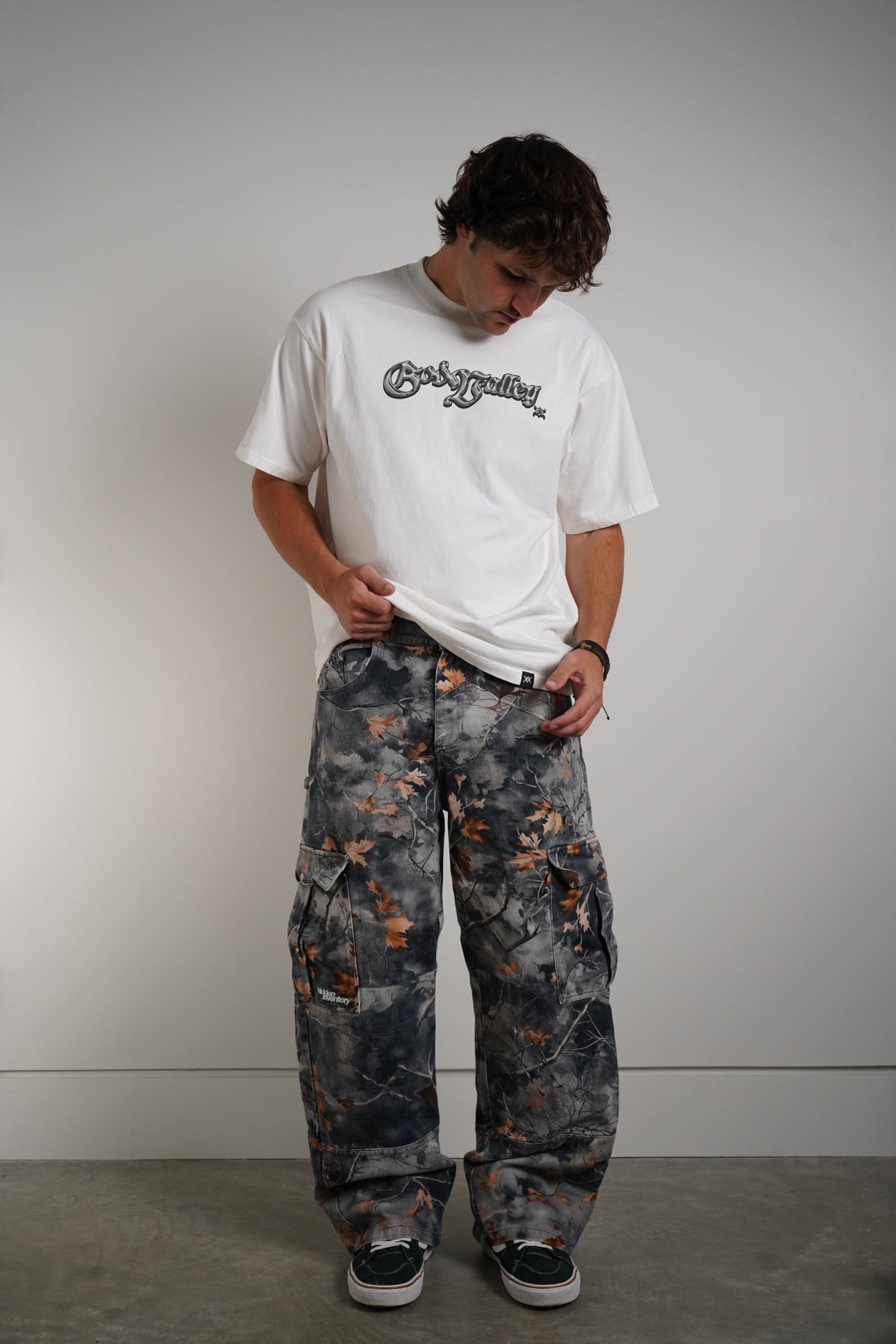 A man wearing a white graphic t-shirt and baggy gray camouflage cargo pants with orange accents, standing against a plain gray wall.