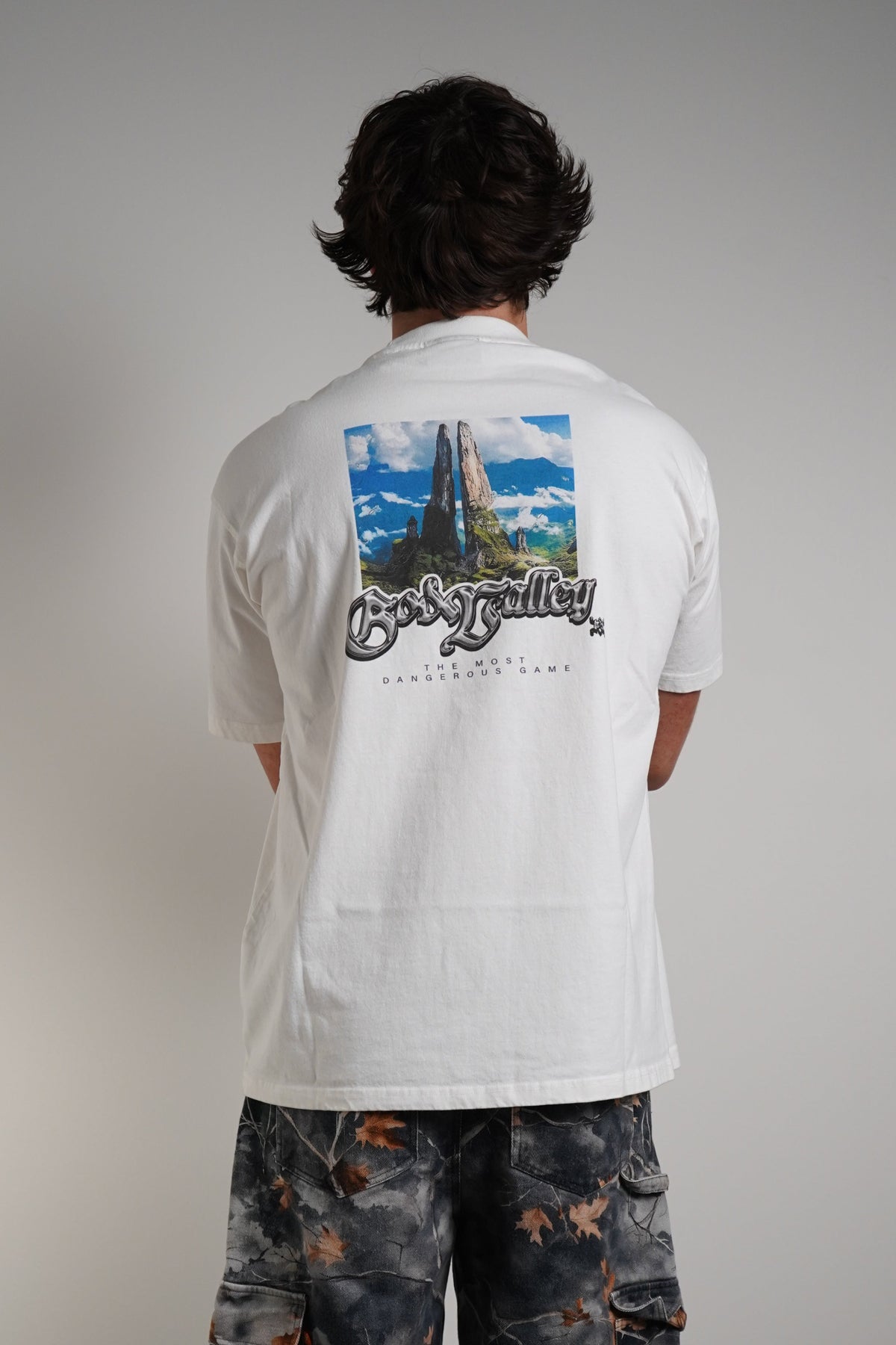 A person shown from the back wearing a white short-sleeve t-shirt with a large graphic print on the back and wide-leg dark camouflage cargo pants, standing against a plain light gray wall.