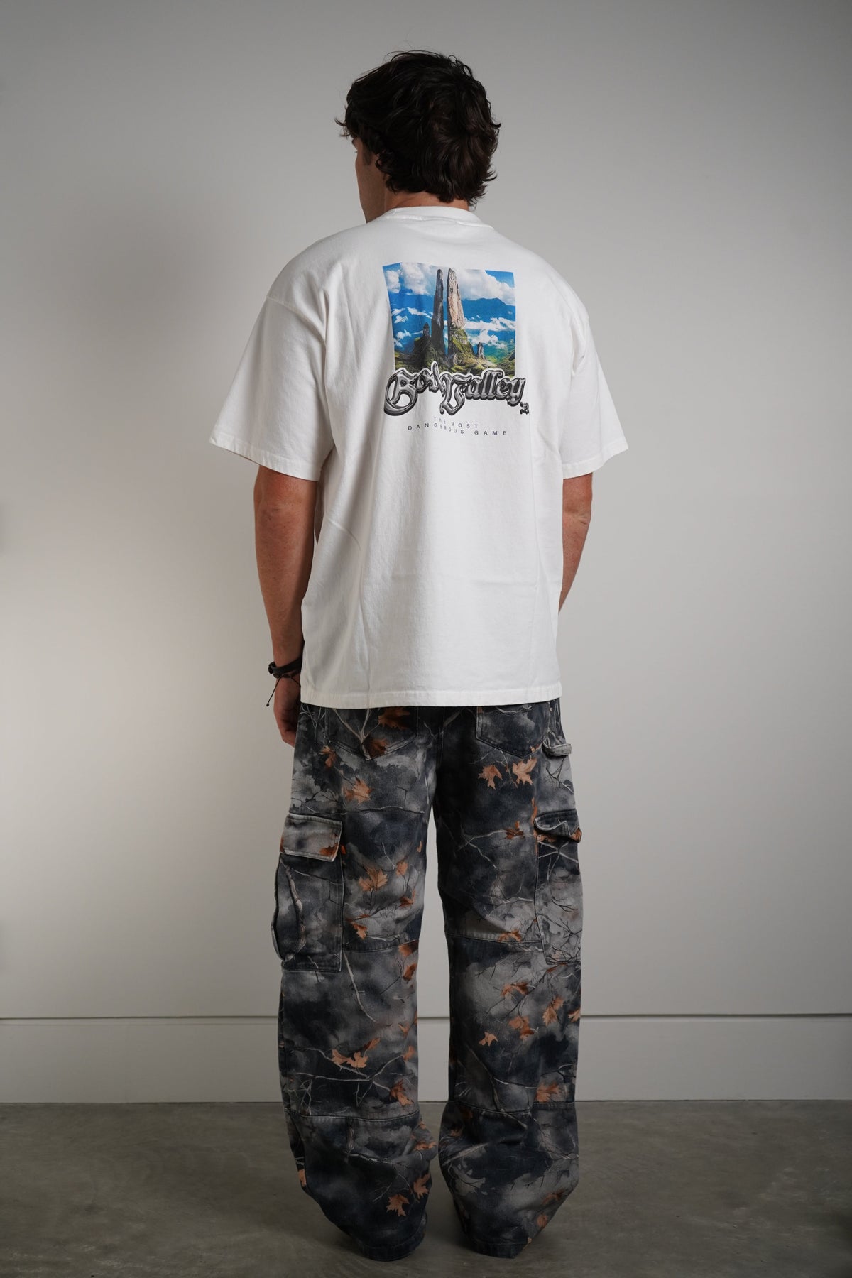 A man stands with his back to the camera wearing a white t-shirt with a large graphic print on the back and wide-leg dark camouflage cargo pants.