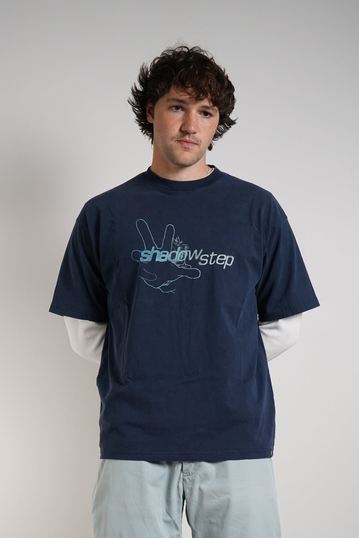 A man wearing a navy blue t-shirt with a light blue graphic print, layered over a white long-sleeve shirt, paired with light blue shorts, white socks, and white sneakers, standing against a plain light gray wall.