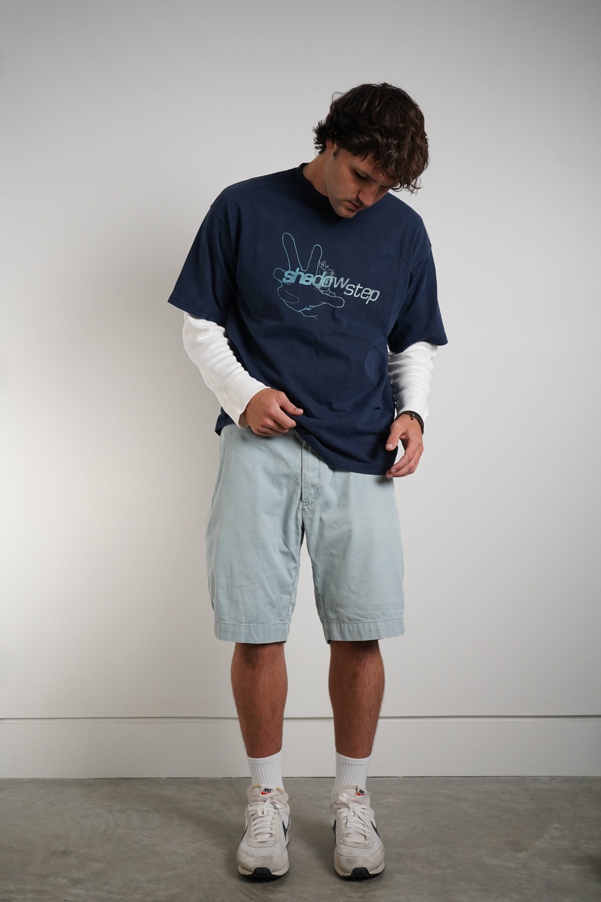 A man wearing a navy blue t-shirt with a 'shadowstep' graphic, layered over a white long-sleeve shirt, paired with light blue shorts, white socks, and white sneakers, standing against a plain light gray wall.