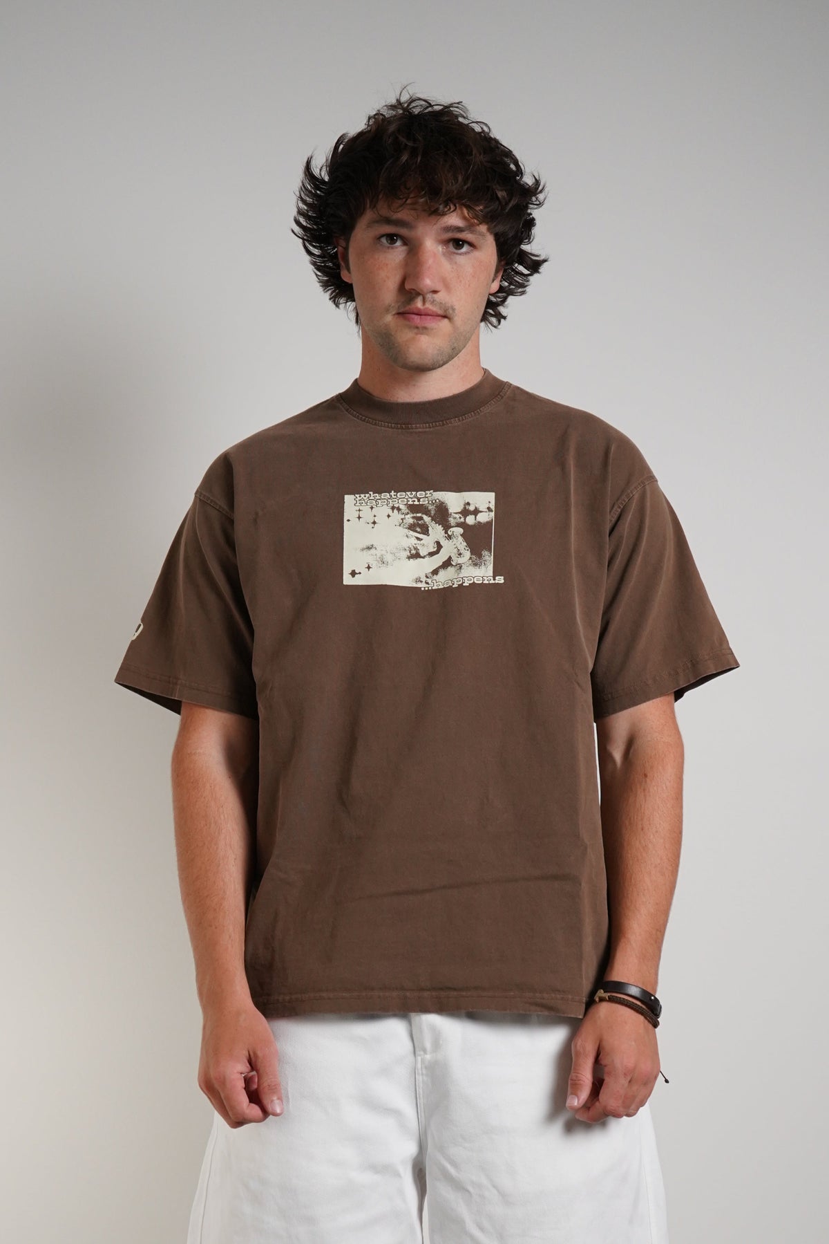 A man stands against a plain light gray wall wearing a brown short-sleeve graphic T-shirt, loose white pants, and black sneakers.