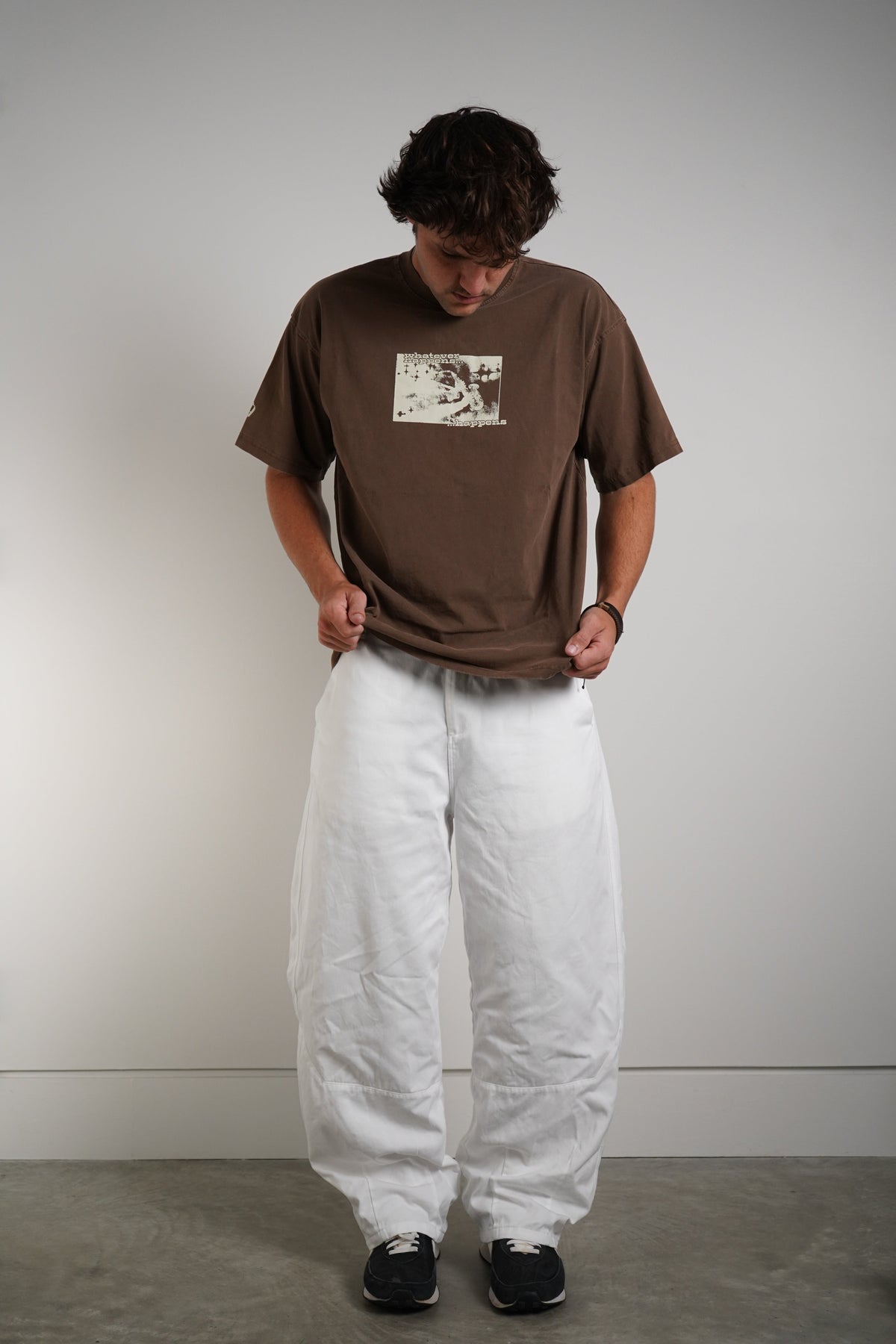 A person standing against a plain light gray wall wearing an oversized brown graphic T-shirt, loose white pants, and black sneakers.