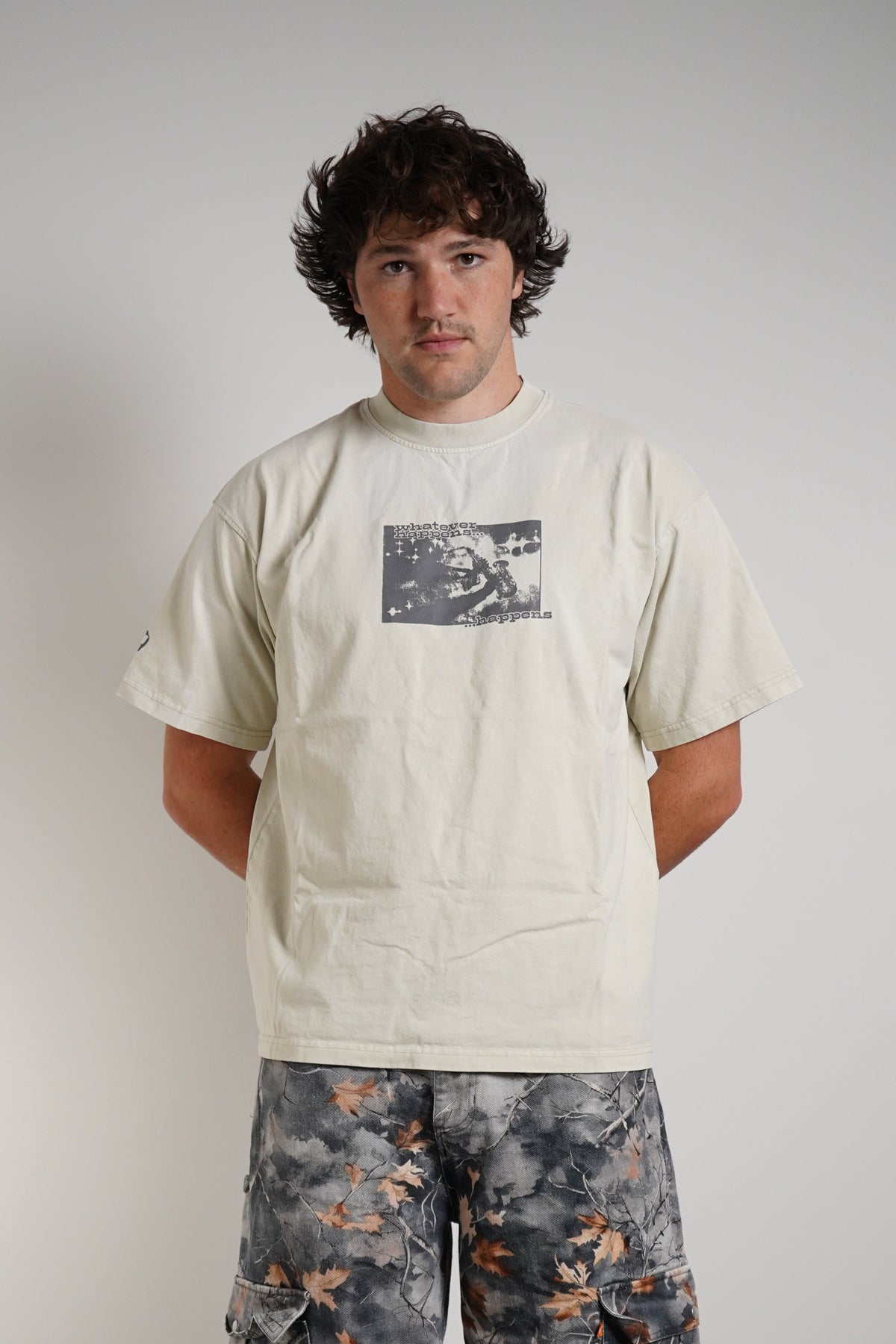 A man stands against a plain light gray wall wearing a cream-colored graphic t-shirt, loose-fitting gray camouflage cargo pants with orange accents, and black boots.