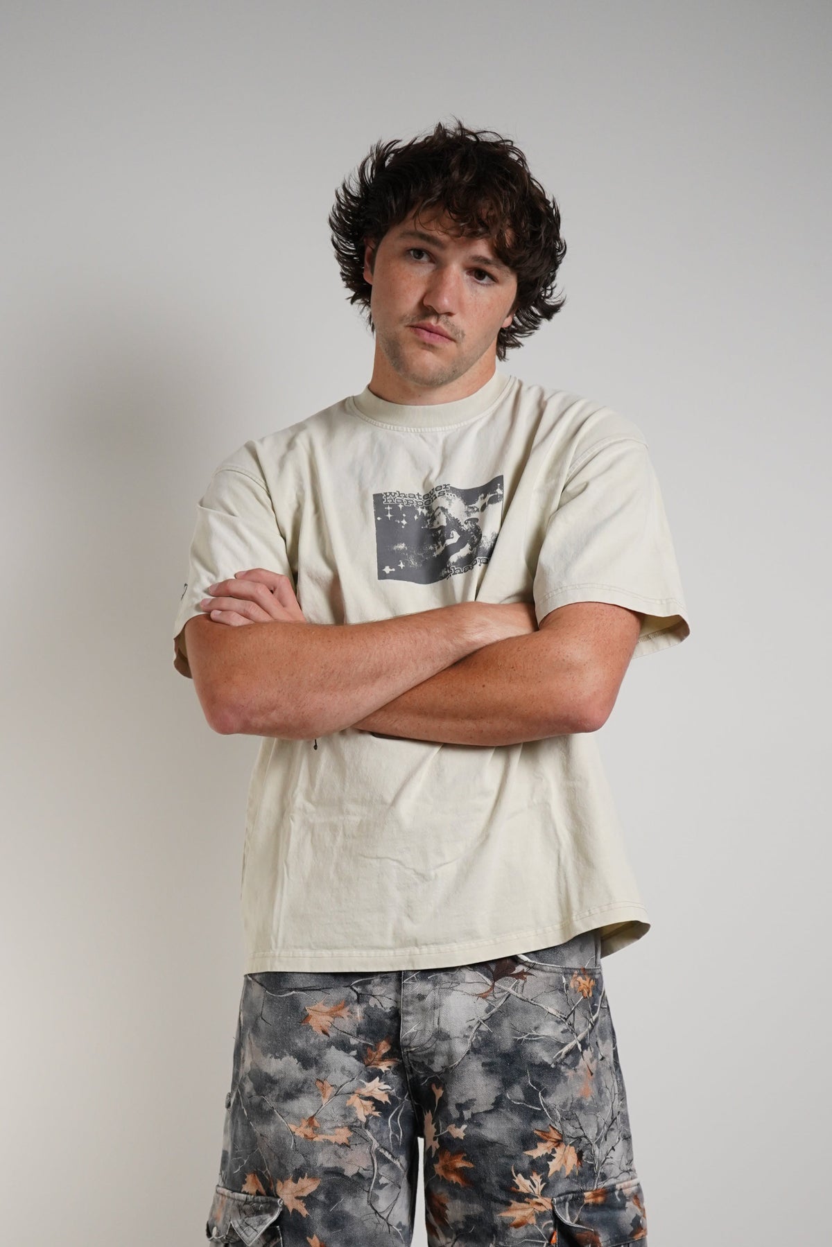 A man stands with arms crossed wearing a cream graphic t-shirt, loose-fit gray camouflage cargo pants with orange accents, and black boots, set against a plain light gray studio background.
