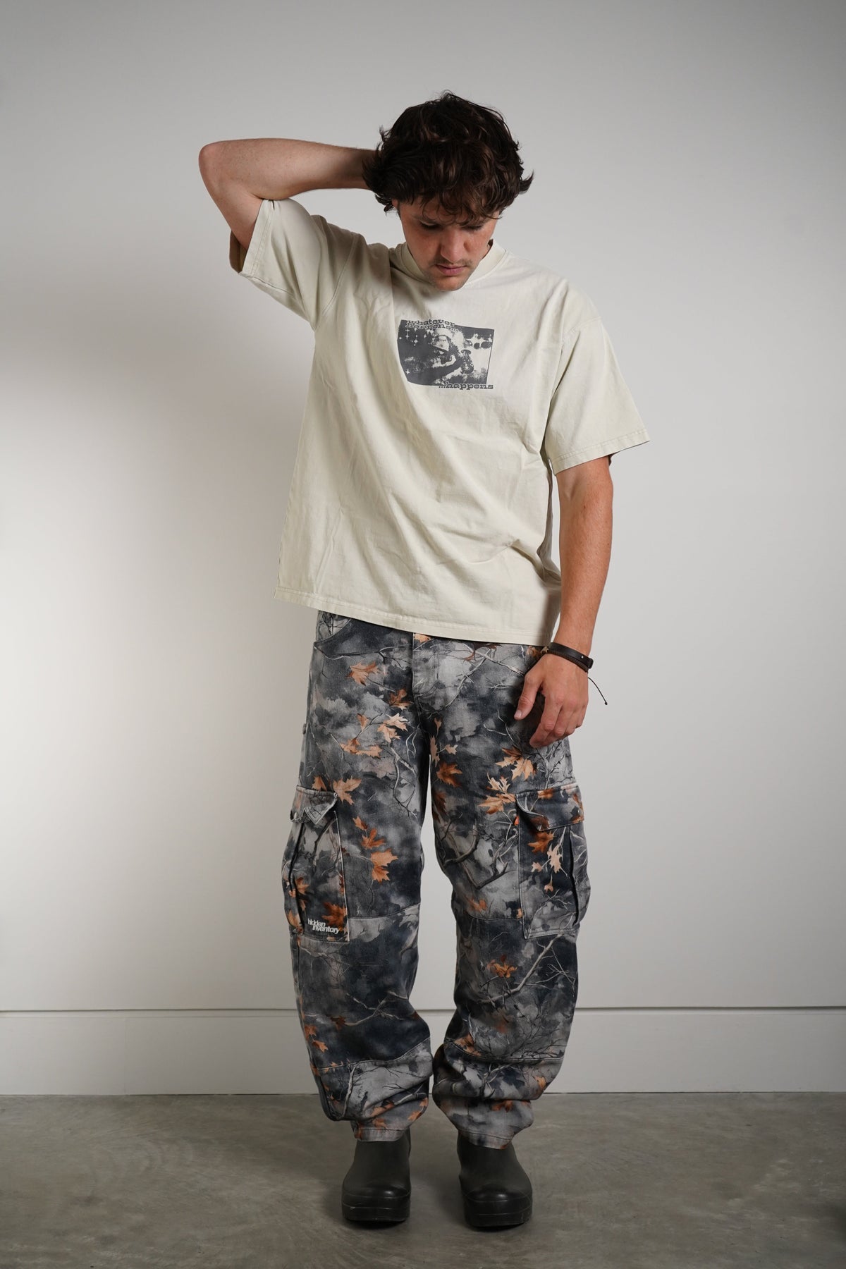 A model stands against a plain light gray wall wearing a cream-colored graphic t-shirt and baggy gray camouflage cargo pants with orange leaf accents.