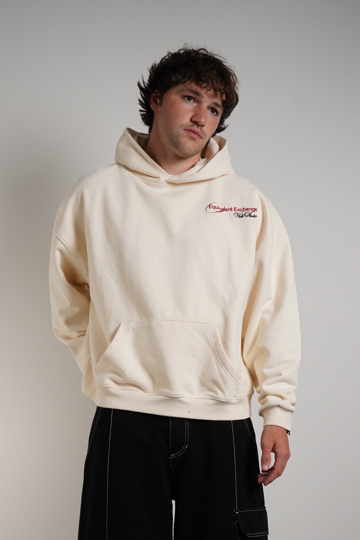 A man wearing a cream-colored hoodie with a small red and black logo on the left chest, paired with black cargo pants, standing against a plain light gray wall.
