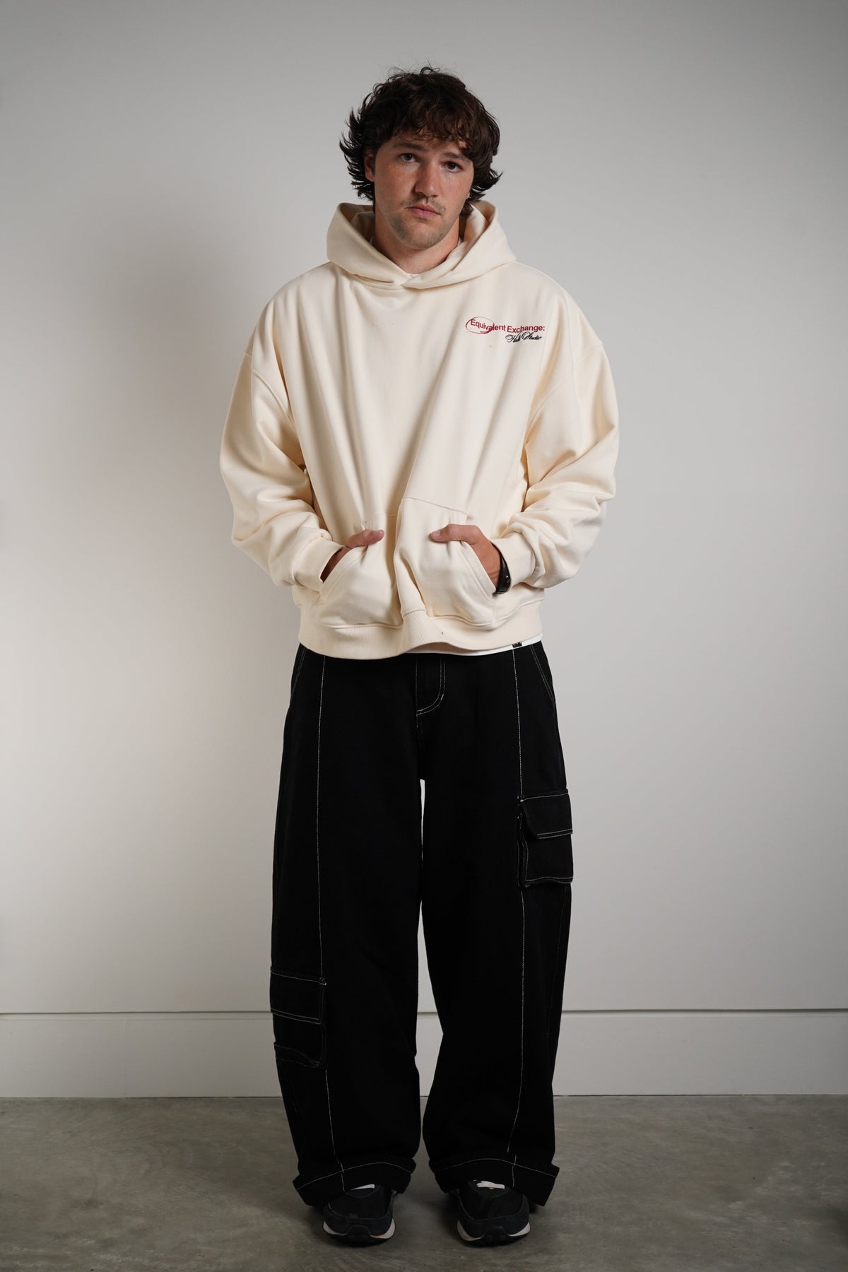 A man wearing a cream-colored hoodie with a small red and black logo on the left chest, paired with black cargo pants and black sneakers, standing against a plain gray wall.