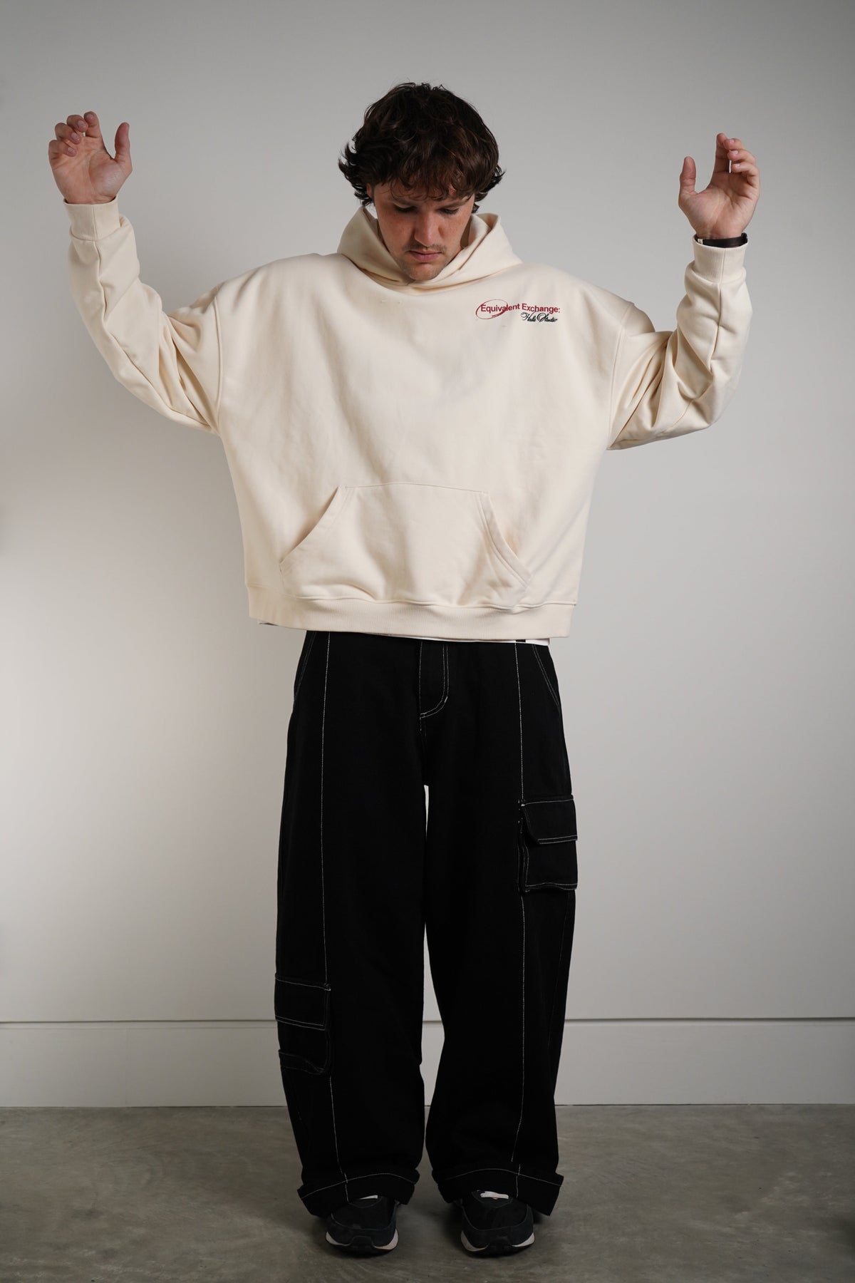 A person wearing a cream-colored cropped hoodie with a small red and blue graphic on the chest, paired with black cargo pants, standing against a plain light gray wall.