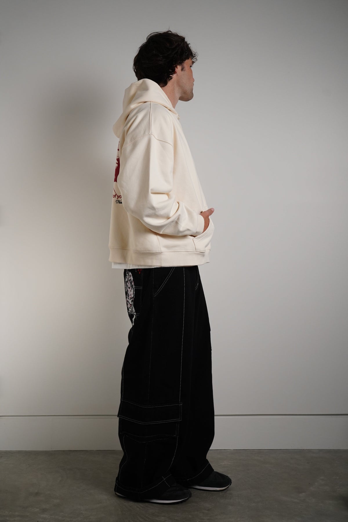 A man stands in profile wearing a cream-colored hoodie with a red graphic on the back and black wide-leg pants, set against a plain gray studio wall.