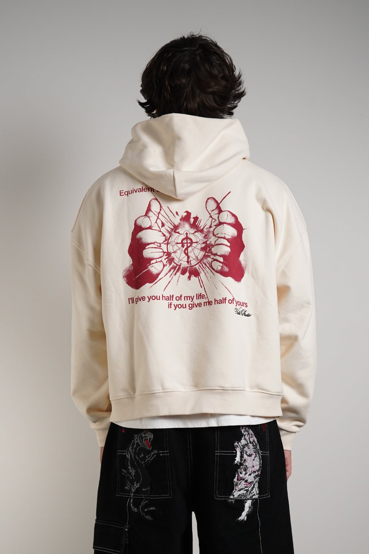 A model shown from the back wearing a cream-colored hoodie with a large red graphic print on the back, paired with black wide-leg pants featuring white graphic details, set against a plain gray studio background.
