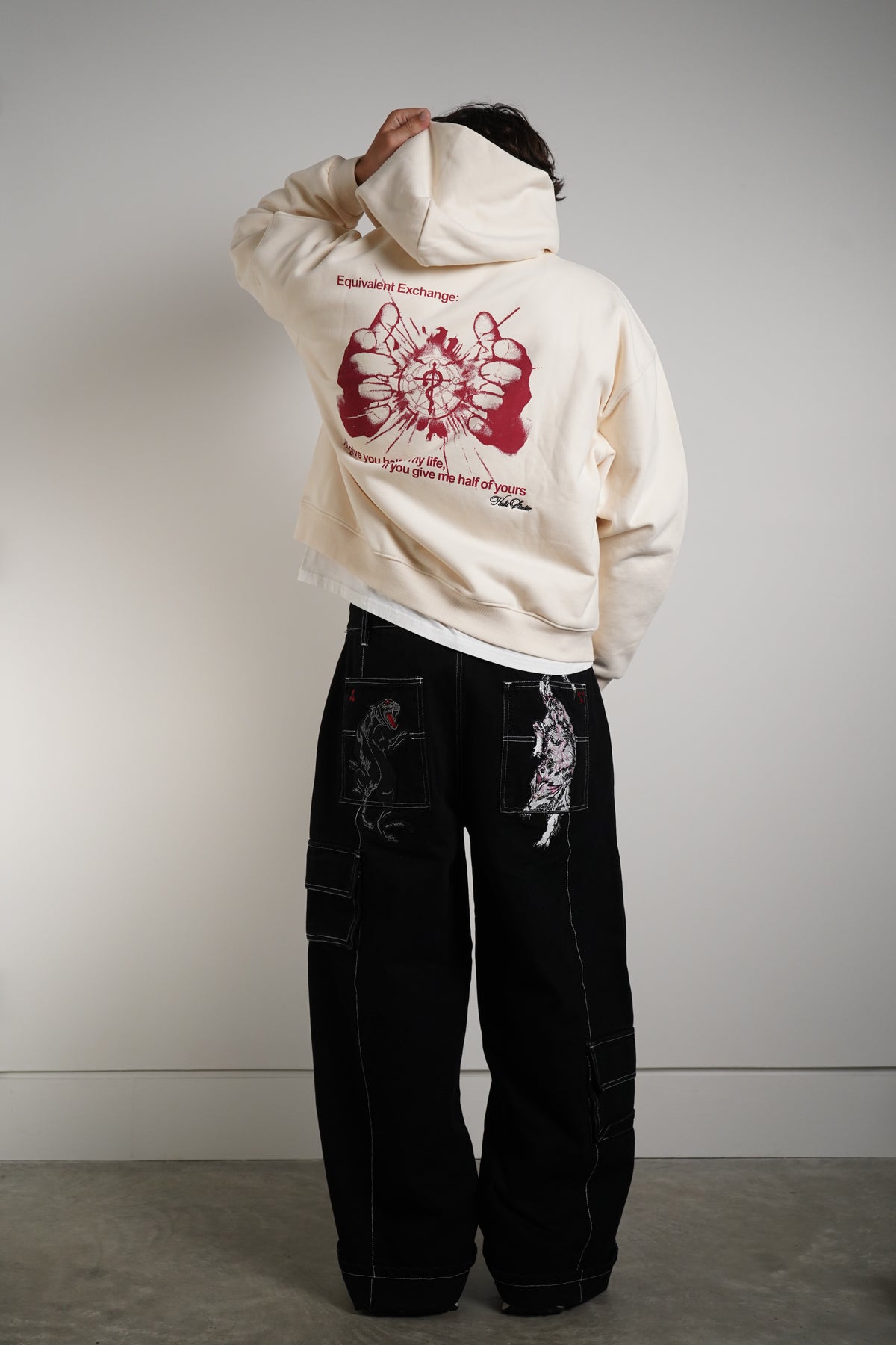 Back view of model in the cream Equivalent Exchange Hoodie with red hands and circle graphic and printed text, worn with black cargo pants