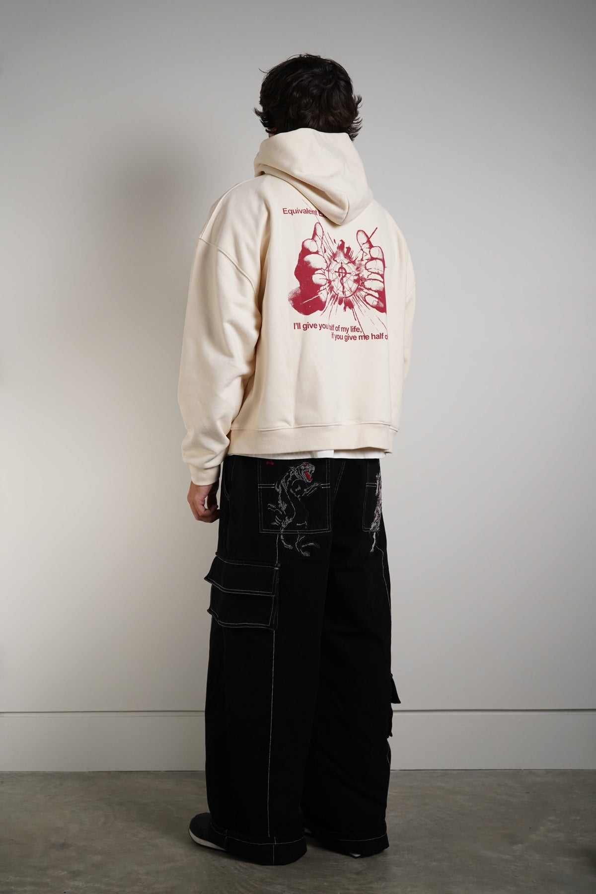 A model shown from the back wearing a cream-colored hoodie with a large red graphic print on the back, paired with black wide-leg cargo pants, in a minimalist studio setting.