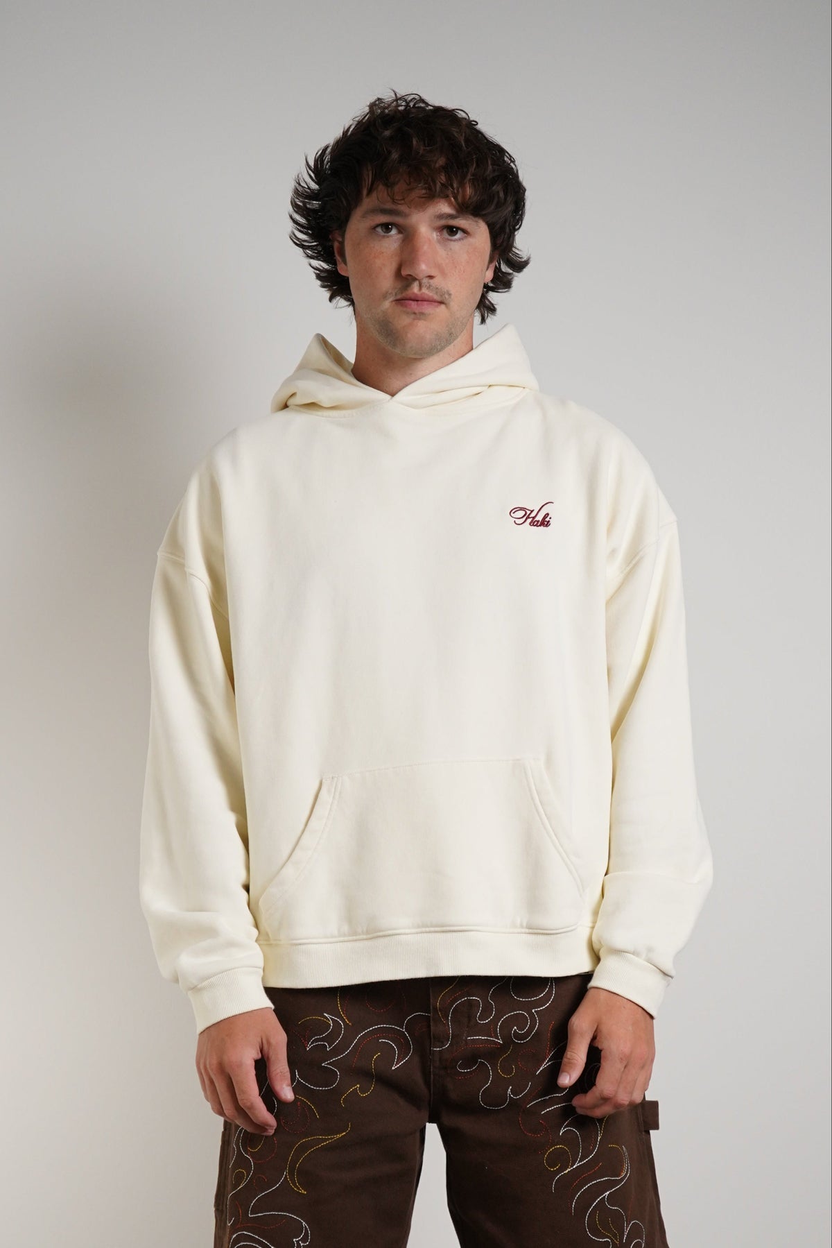 A man stands against a plain light gray wall wearing a cream-colored hoodie with a small red embroidered logo on the left chest, dark brown shorts with a light brown paisley pattern, white socks, and white sneakers.