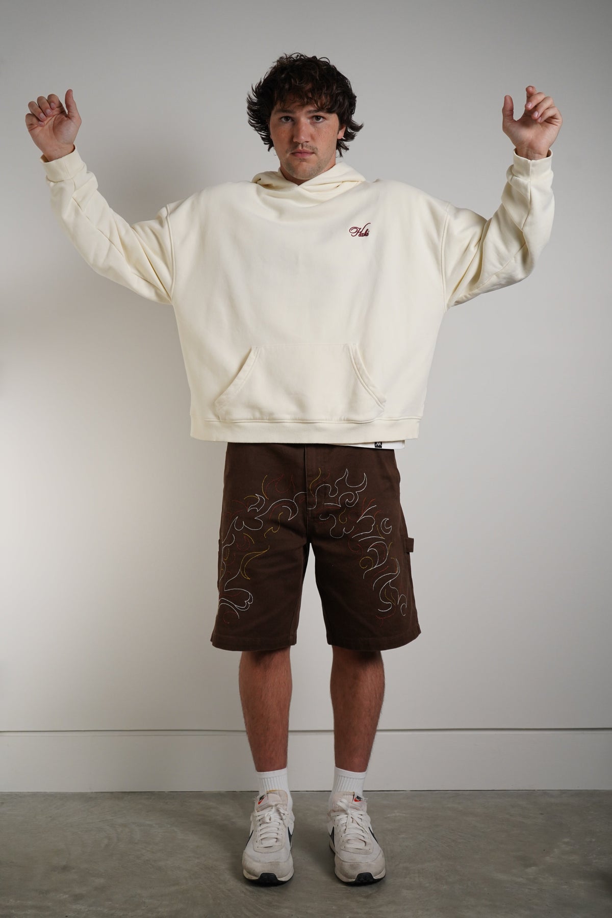A man modeling a cream-colored cropped hoodie with a small red logo on the chest, paired with brown shorts featuring white flame-like patterns, white socks, and white sneakers, standing against a plain light gray wall.