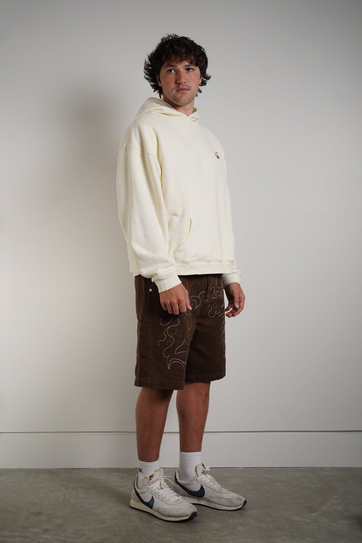 A man stands in profile wearing a cream-colored hoodie, brown shorts with white embroidered details, white socks, and white Nike sneakers against a plain light gray wall.