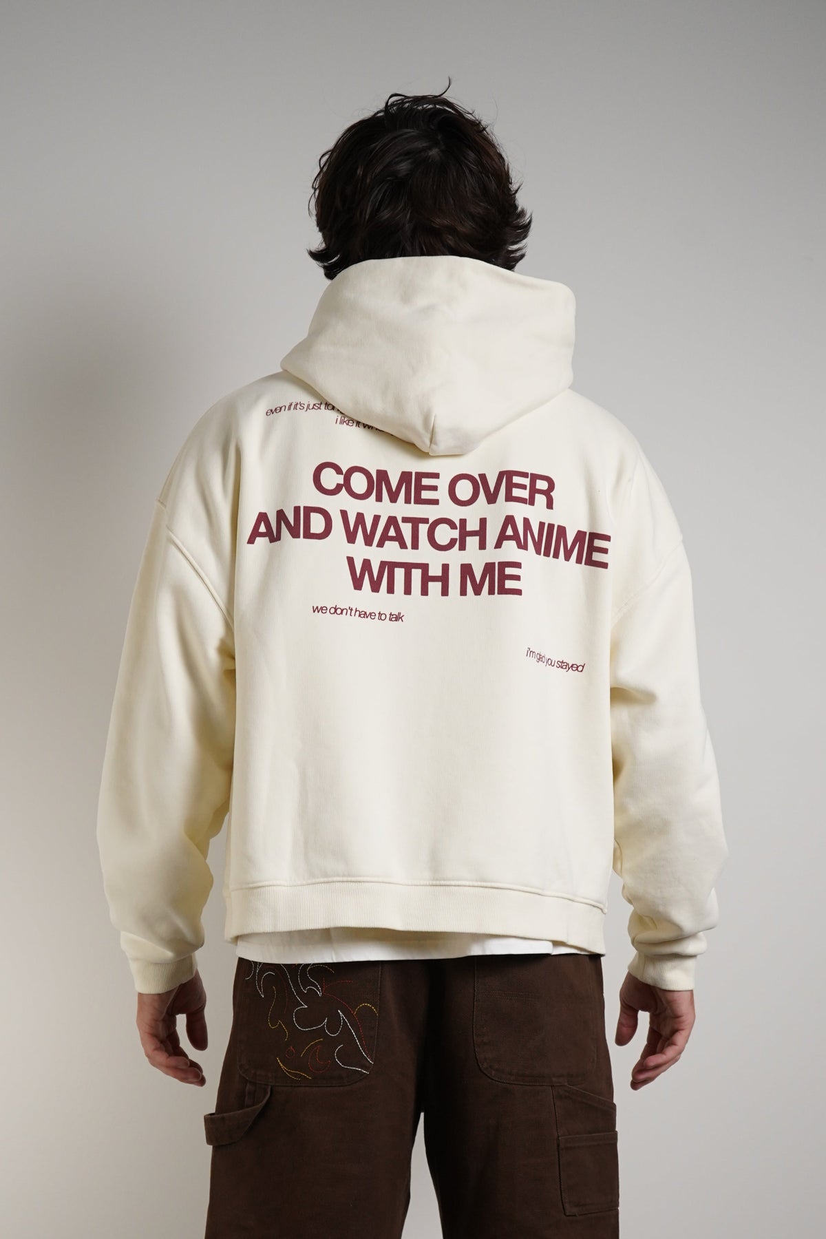A person shown from the back wearing a cream-colored hoodie with red text on the back reading 'COME OVER AND WATCH ANIME WITH ME', paired with brown shorts, white socks, and sneakers, standing against a plain light gray wall.