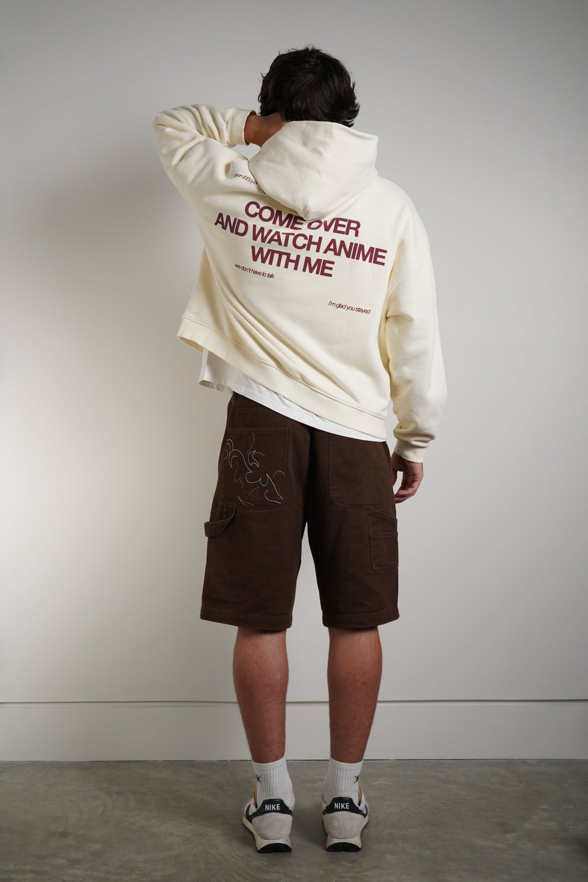 A person shown from behind wearing a cream-colored hoodie with red text on the back, brown cargo shorts, white socks, and sneakers, standing against a plain light gray wall.