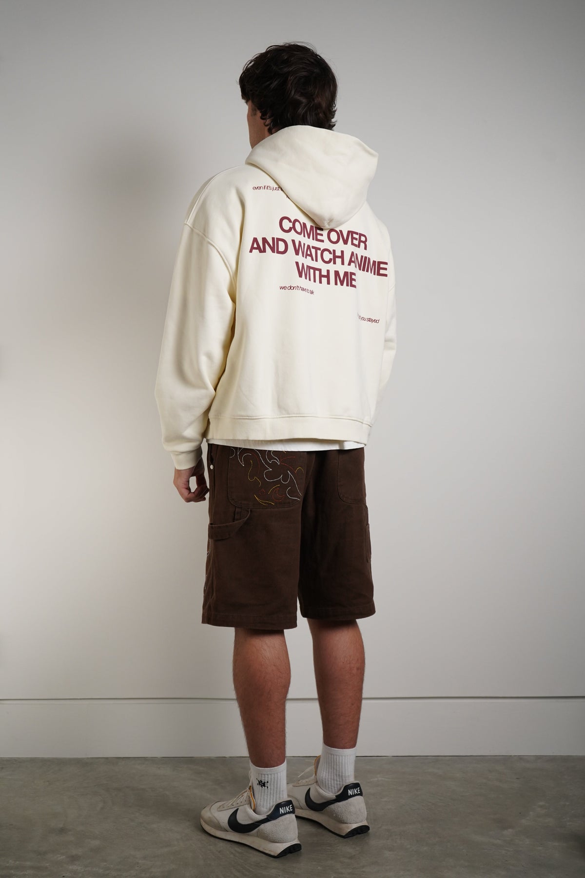 A model shown from the back wearing a cream-colored hoodie with red text on the back reading 'COME OVER AND WATCH ME WITH ME', paired with brown shorts, white socks, and white sneakers with black accents, standing against a plain light gray wall.
