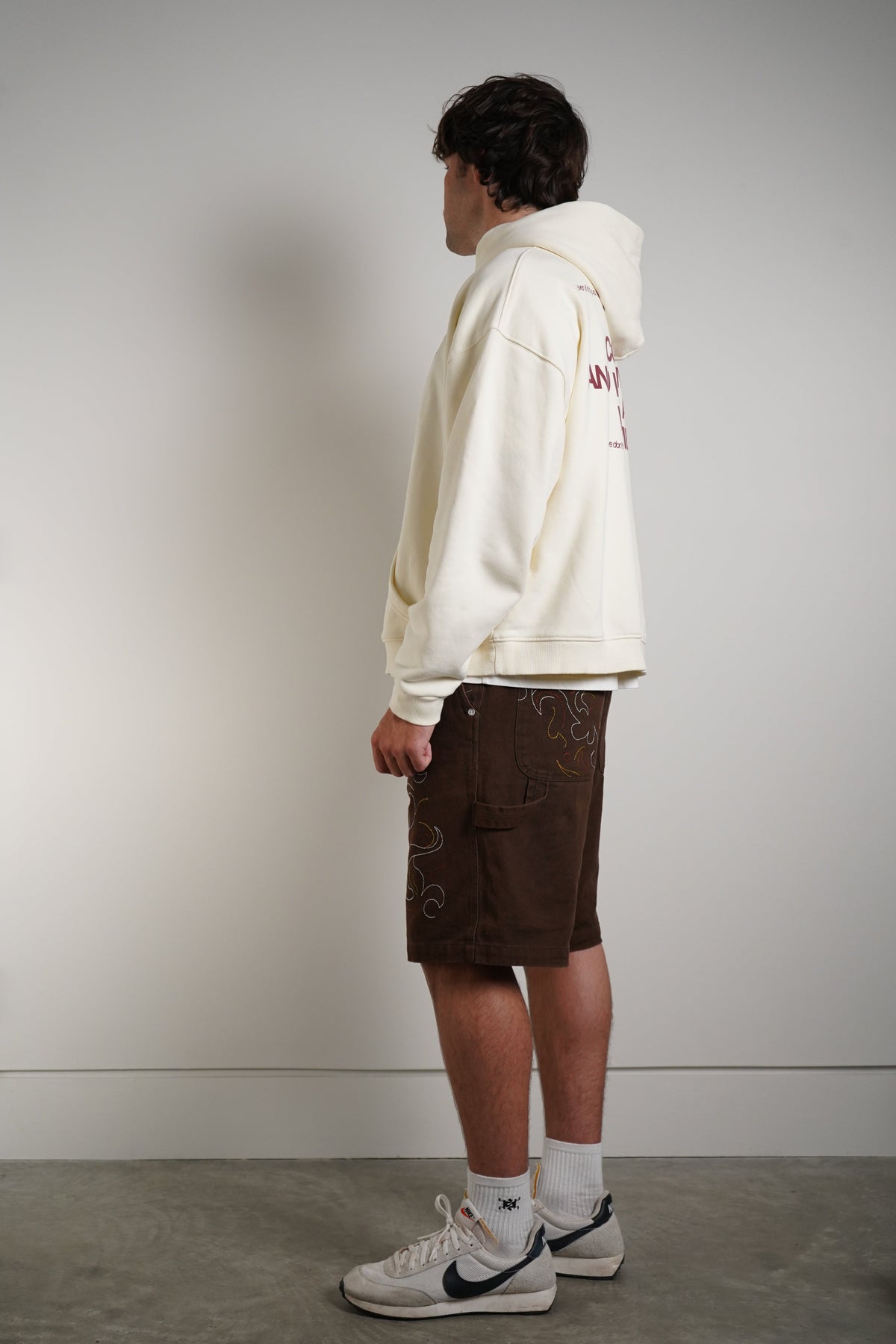 A model standing in profile wearing an oversized cream hoodie with red graphic text on the back, dark brown shorts with subtle gold swirl patterns, white socks, and white Nike Air Force 1 sneakers against a plain light gray wall.