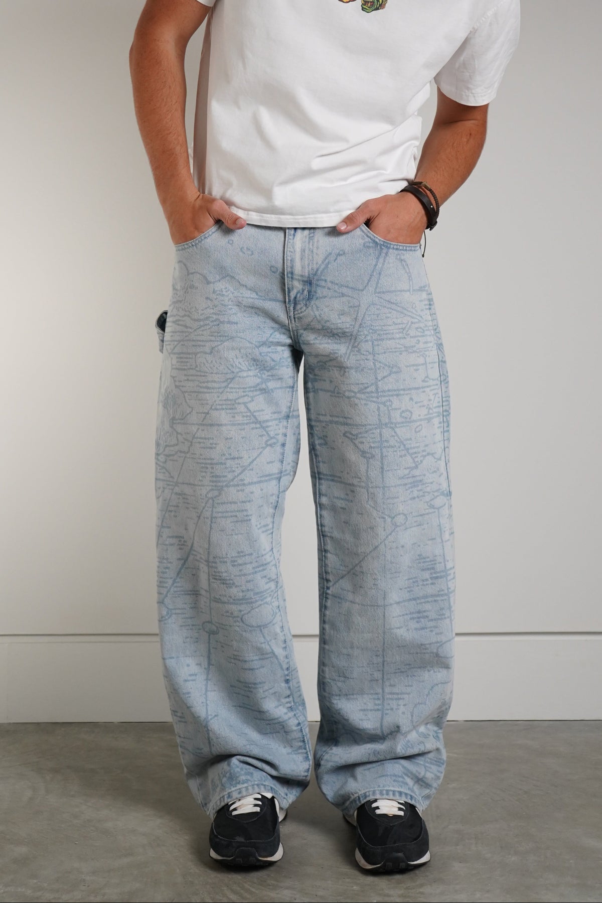 A man wearing a white graphic t-shirt, light-wash wide-leg denim jeans, and black sneakers, standing against a neutral gray studio background.