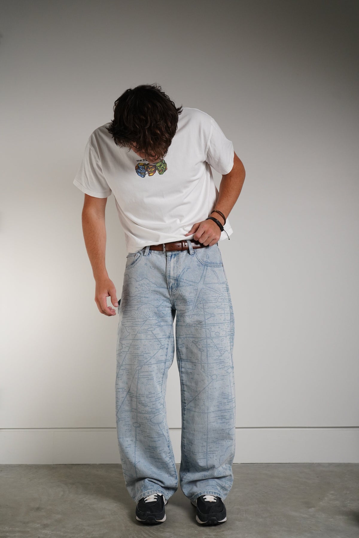 A person wearing a white graphic t-shirt, light-wash wide-leg denim jeans, a brown leather belt, and black sneakers, standing against a neutral gray studio background.