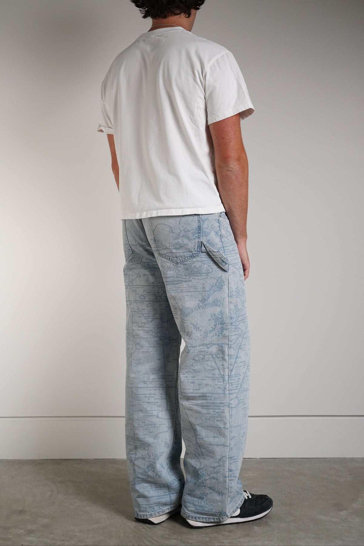 Grand Line Pants in light blue washed denim with an all-over tonal map-style print, worn by a model in a white tee, back three-quarter view