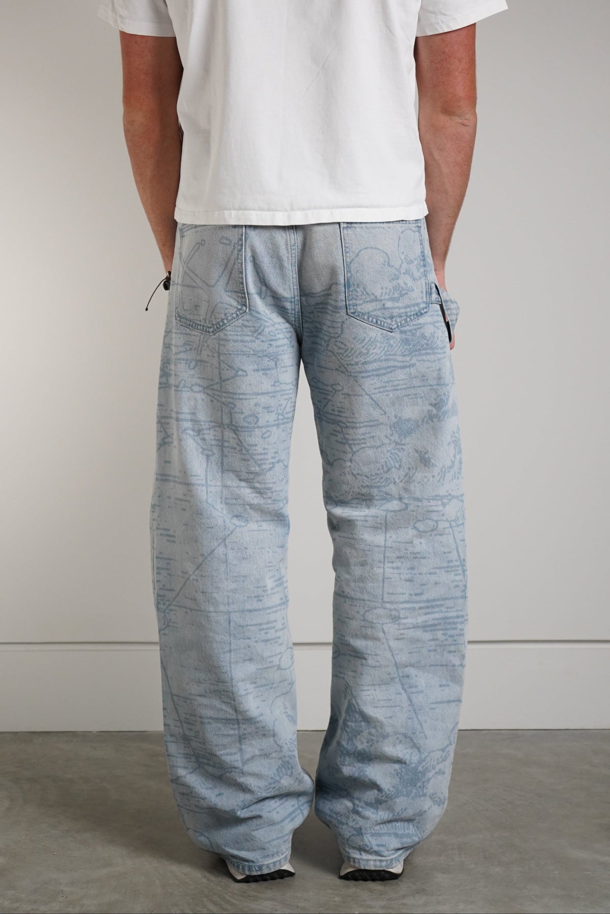 A man viewed from behind wearing a white short-sleeve t-shirt and light blue wide-leg linen pants, standing against a neutral gray studio background.