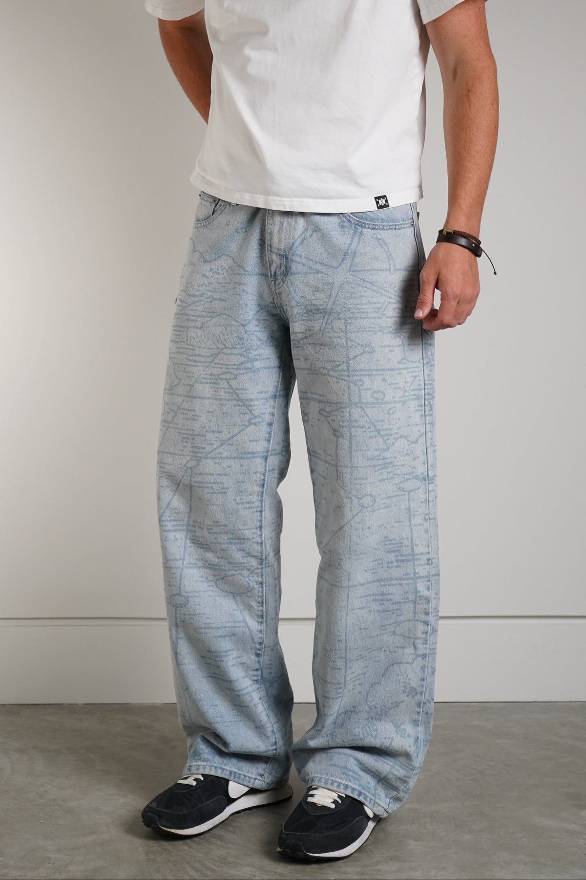 A man wearing a white crewneck t-shirt with a small graphic on the chest, paired with light blue wide-leg jeans and black sneakers, standing against a neutral gray studio background.