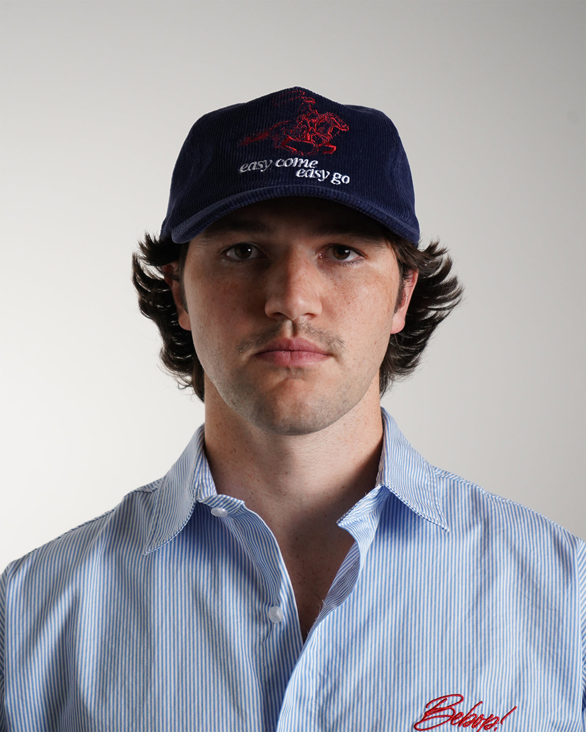 Front view on model of the Easy Come Easy Go Hat in Midnight Blue corduroy, with a red embroidered horse and rider above white embroidered text, worn with a blue striped shirt