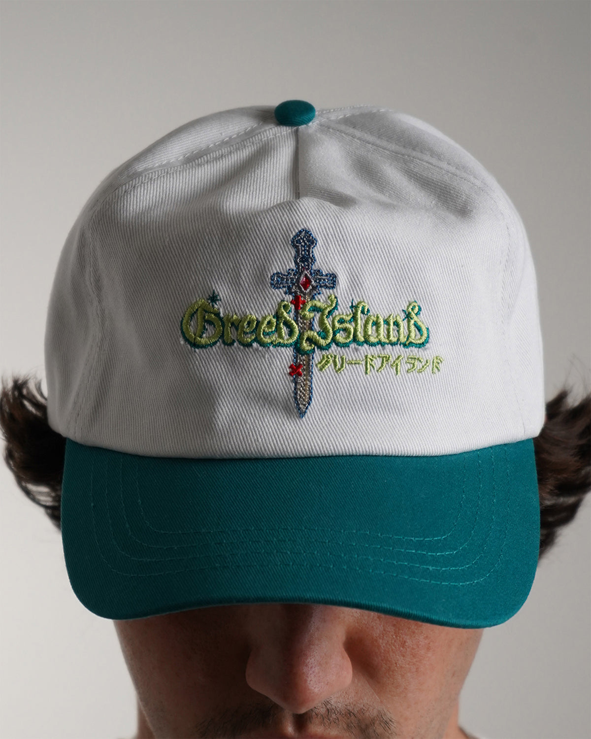 Front view on model of the Greed Island Hat, a white cap with teal brim and button, embroidered sword and green Greed Island lettering with Japanese katakana