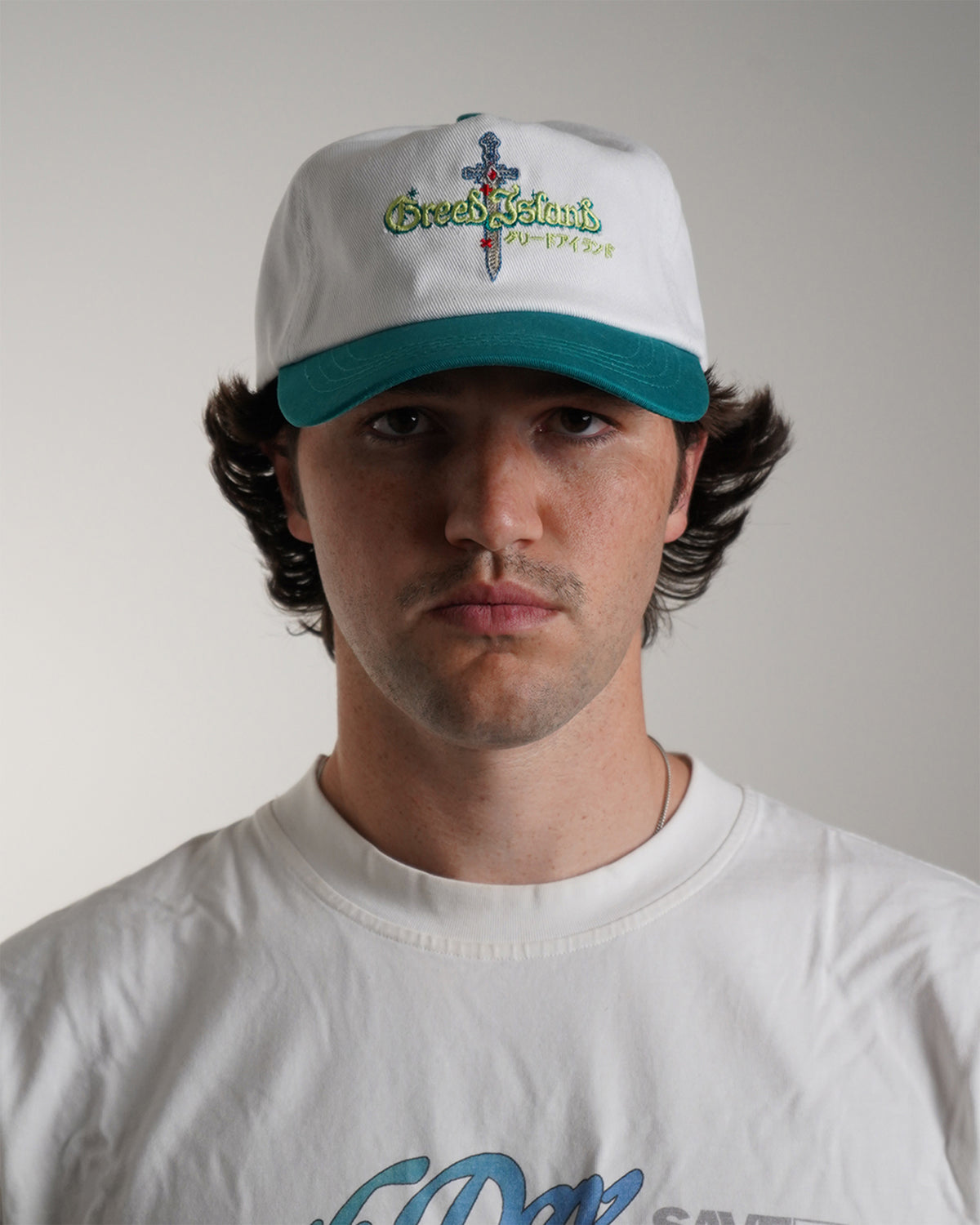 Model wearing the white and teal Greed Island Hat with embroidered dagger logo, front view
