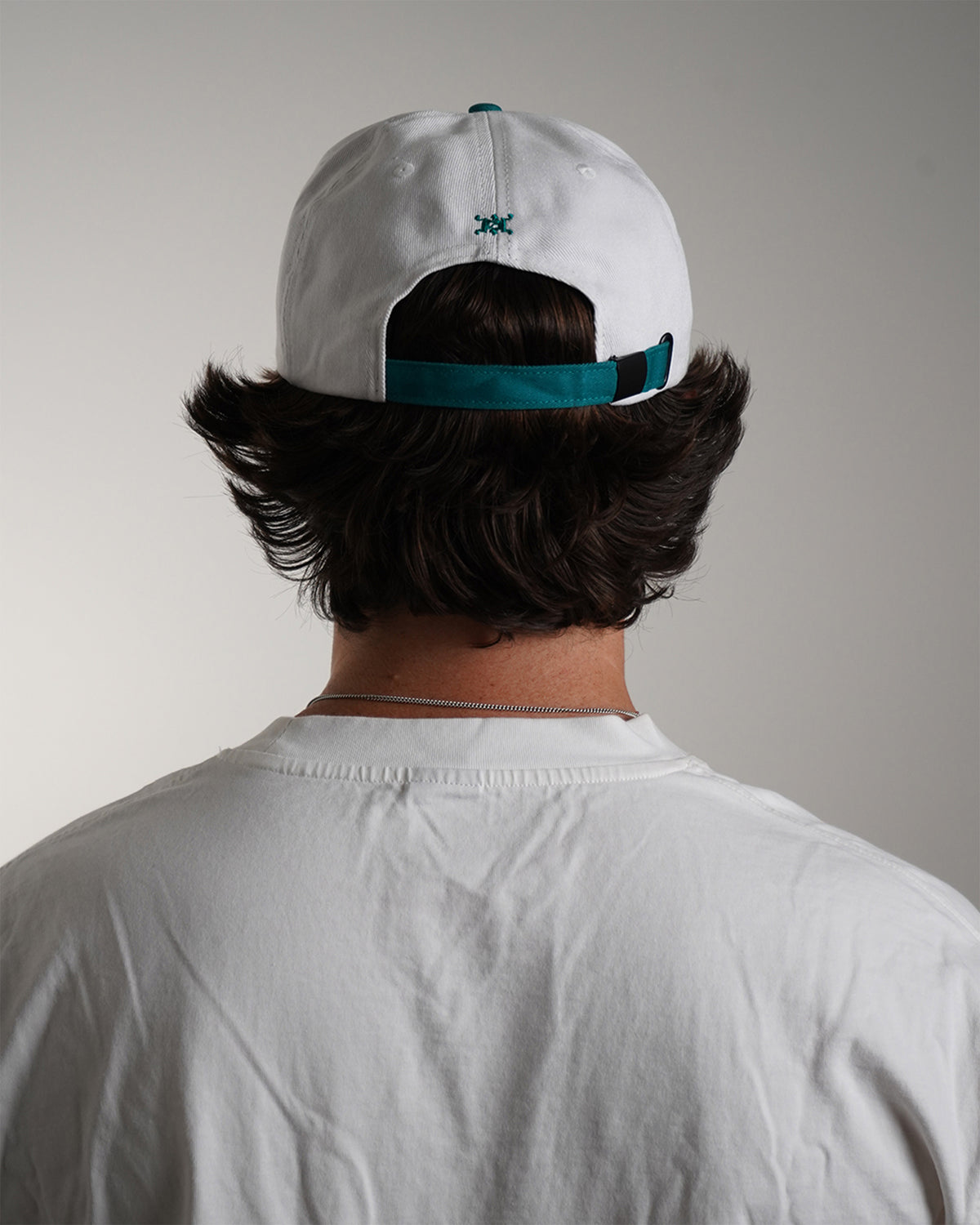 Model wearing the white and teal Greed Island Hat with embroidered haki logo, back view