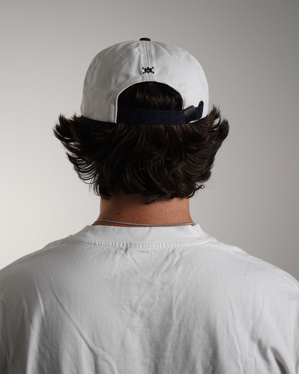 Modeled back view of the white Superrrr Hat showing navy adjustable strap and small black embroidered logo