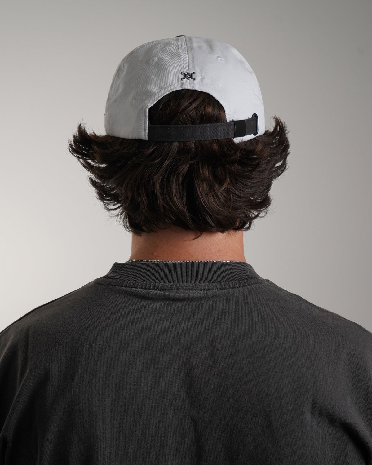 Back view on model of the white Fire Fist Hat with black adjustable strap and small dark embroidered emblem, worn with a dark gray tee
