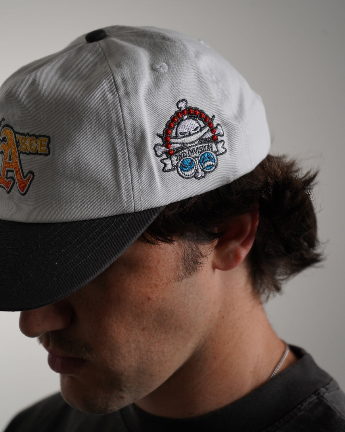 Side close-up on model of the white Fire Fist Hat with black brim and top button, showing a small embroidered skull emblem with red beads and "2ND DIVISION" banner